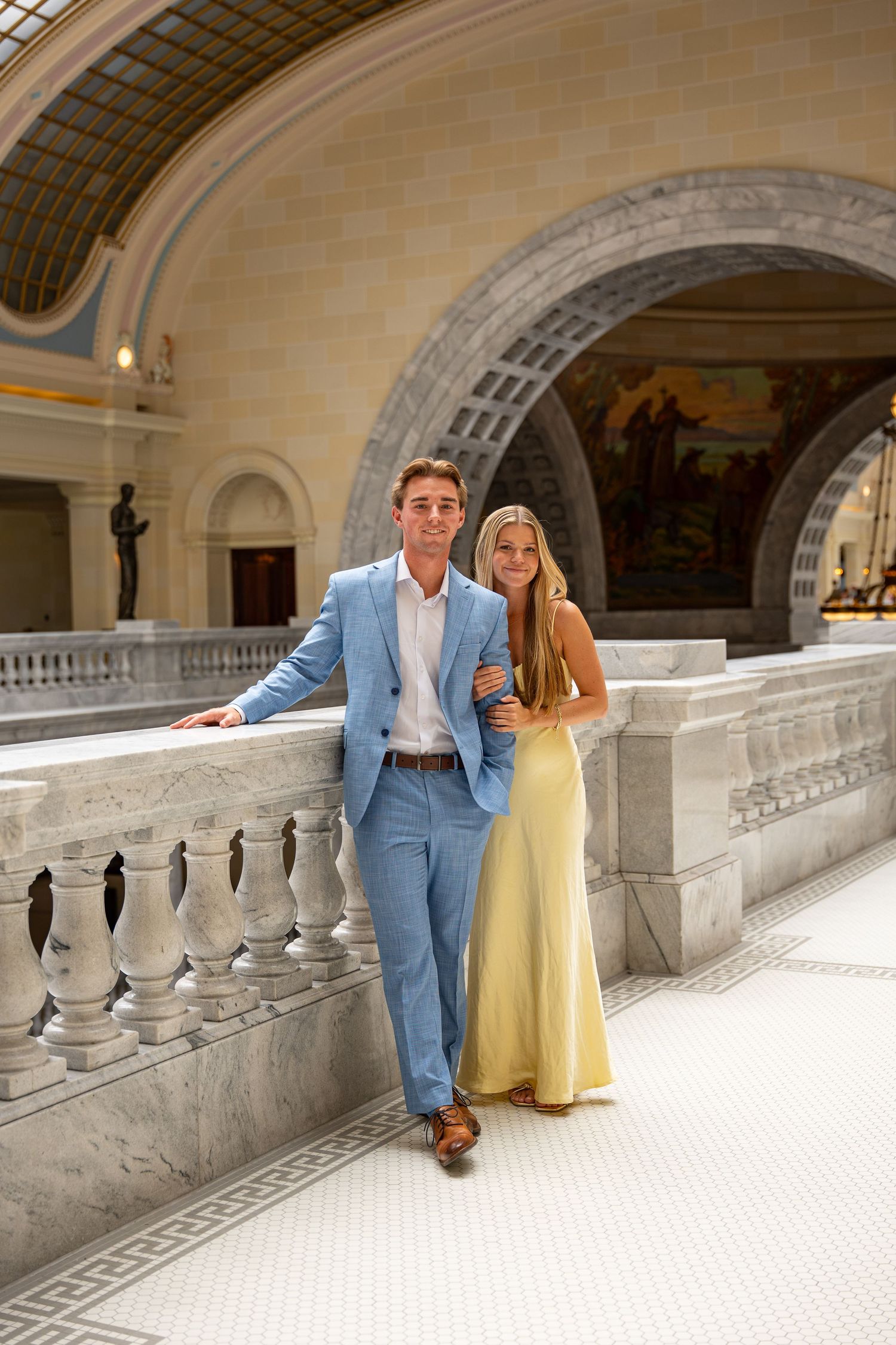 Elegant couple posing at marble balustrade in ornate neoclassical building, man in blue suit, woman in yellow gown.