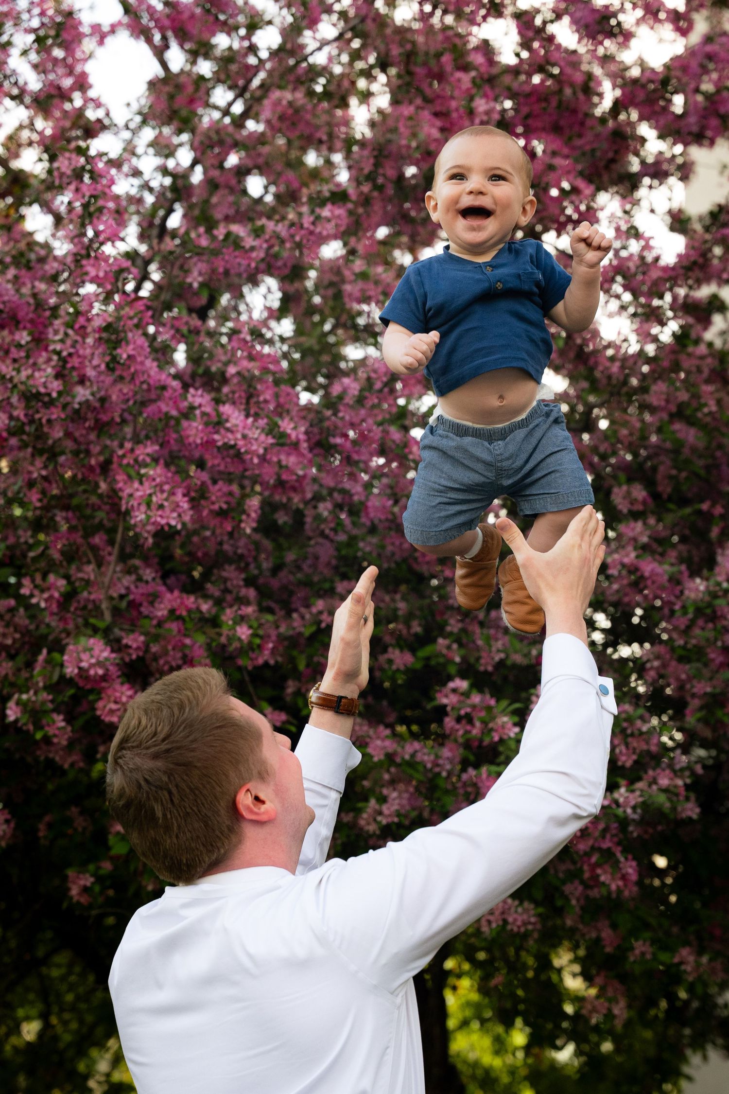 A man holds a young child up in the air in front of blooming trees.