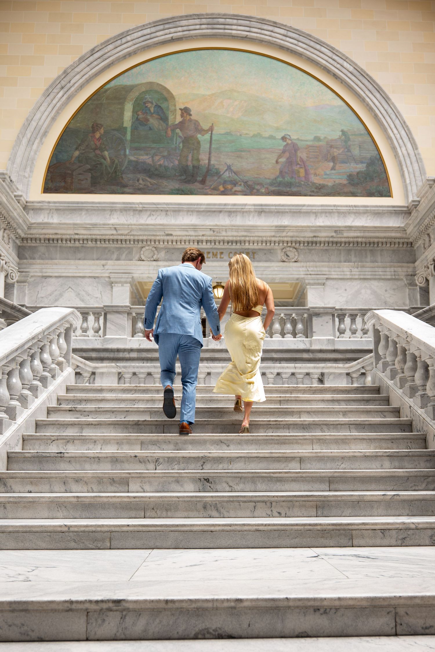 Couple in formal attire running up marble stairs in elegant building with mural.