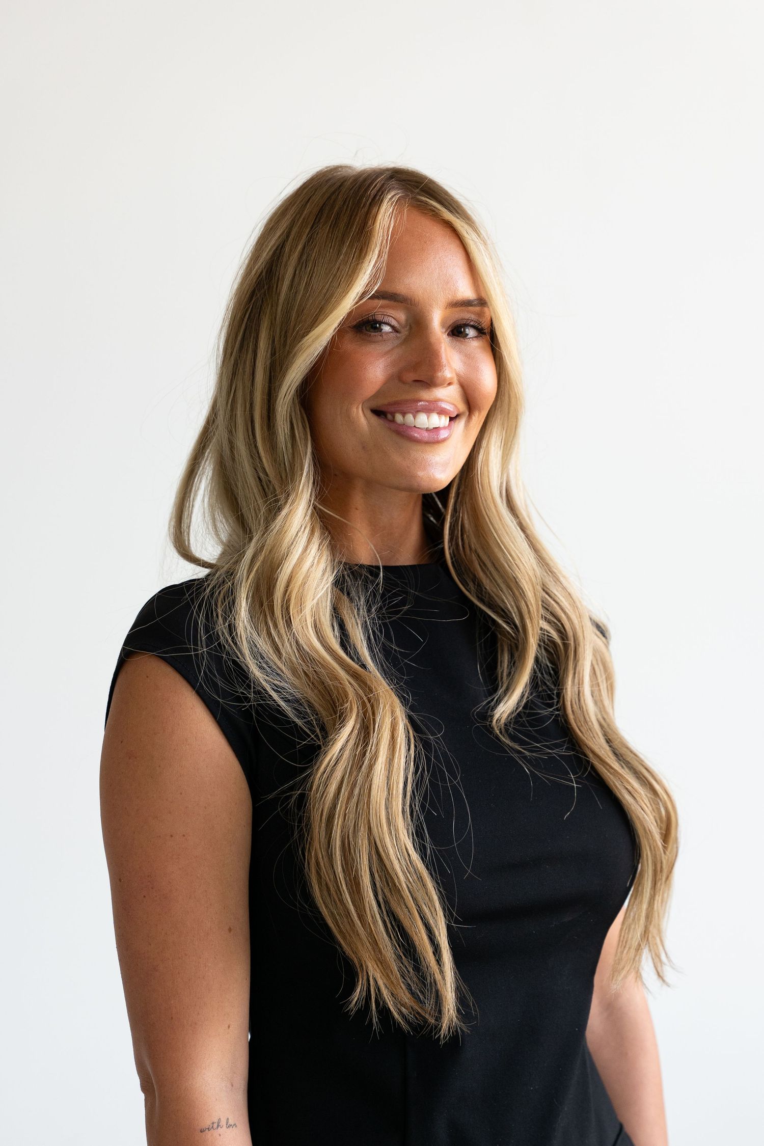 Smiling woman with long blonde wavy hair wearing a black sleeveless top against a white background.