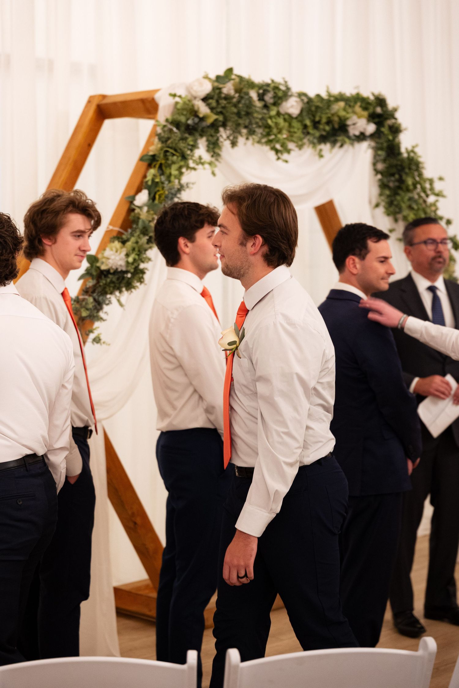Groomsmen in white shirts and orange ties stand near a hexagonal wooden arch decorated with greenery at a wedding.