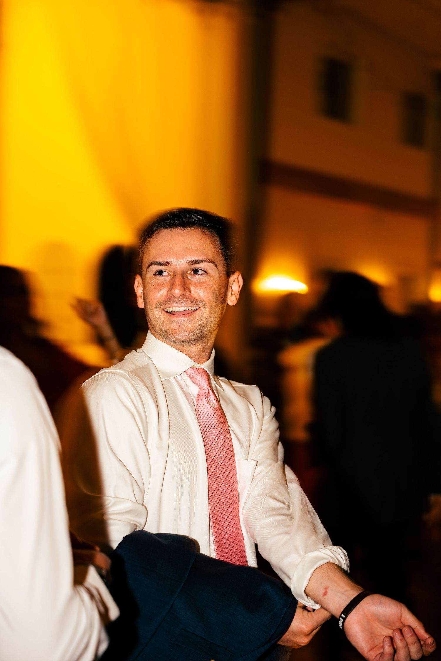 Smiling man in white shirt and pink tie seated at warm golden-lit wedding reception event.