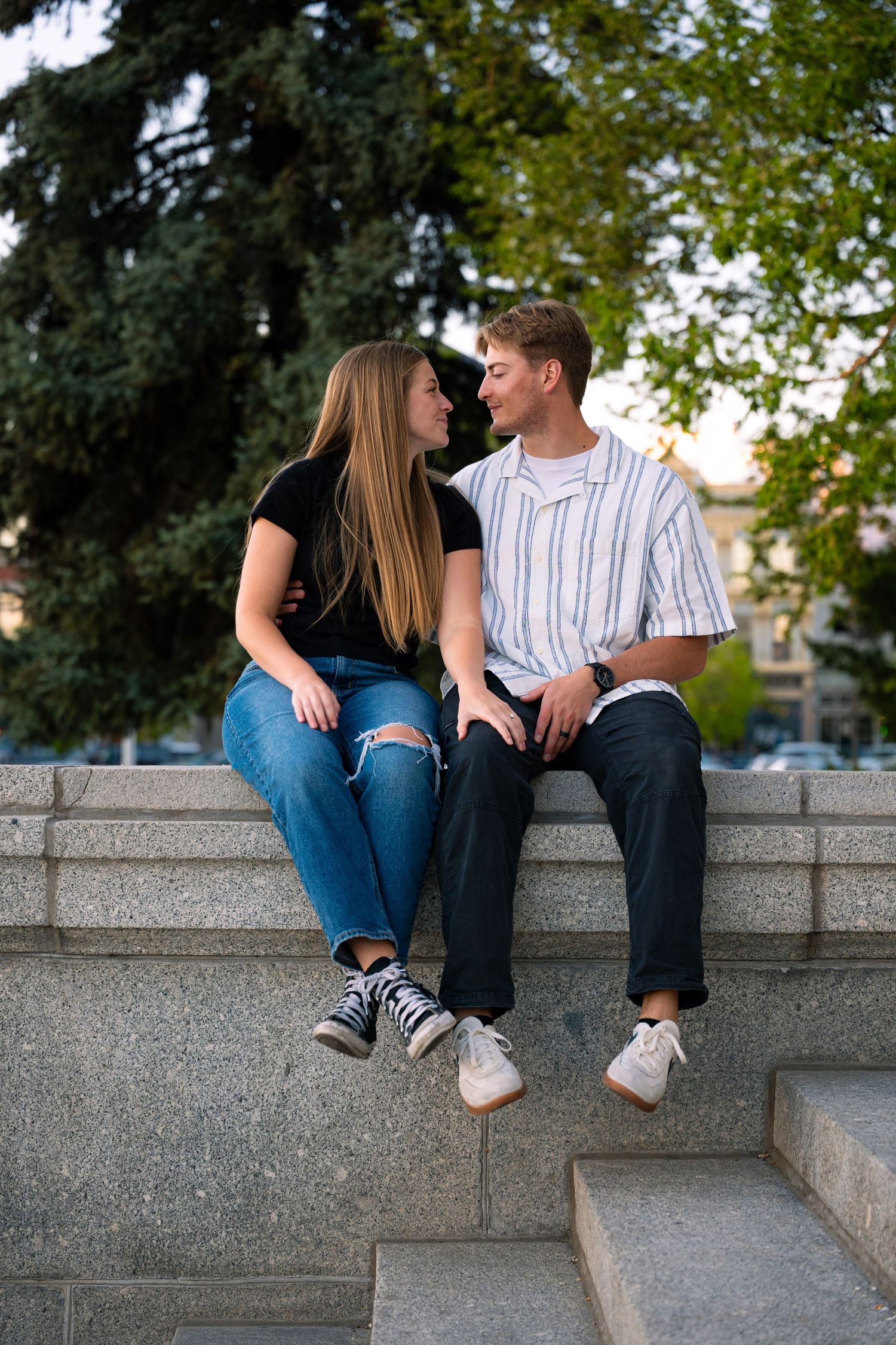 Young couple sitting close on stone steps in a park, sharing a romantic moment surrounded by green trees.