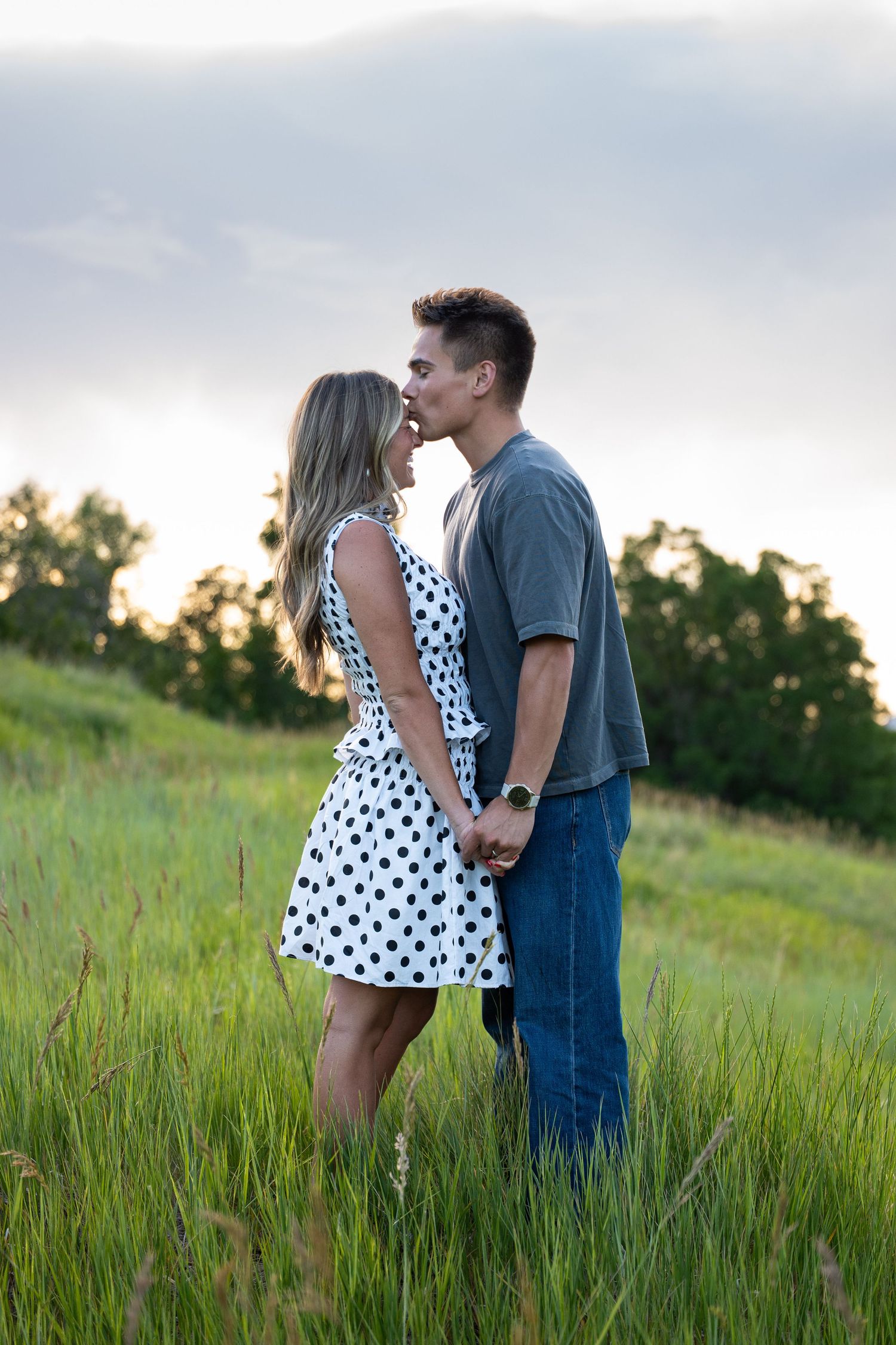 A couple shares a romantic kiss in a lush green field at sunset, woman in polka dot dress holding hands with man in jeans.