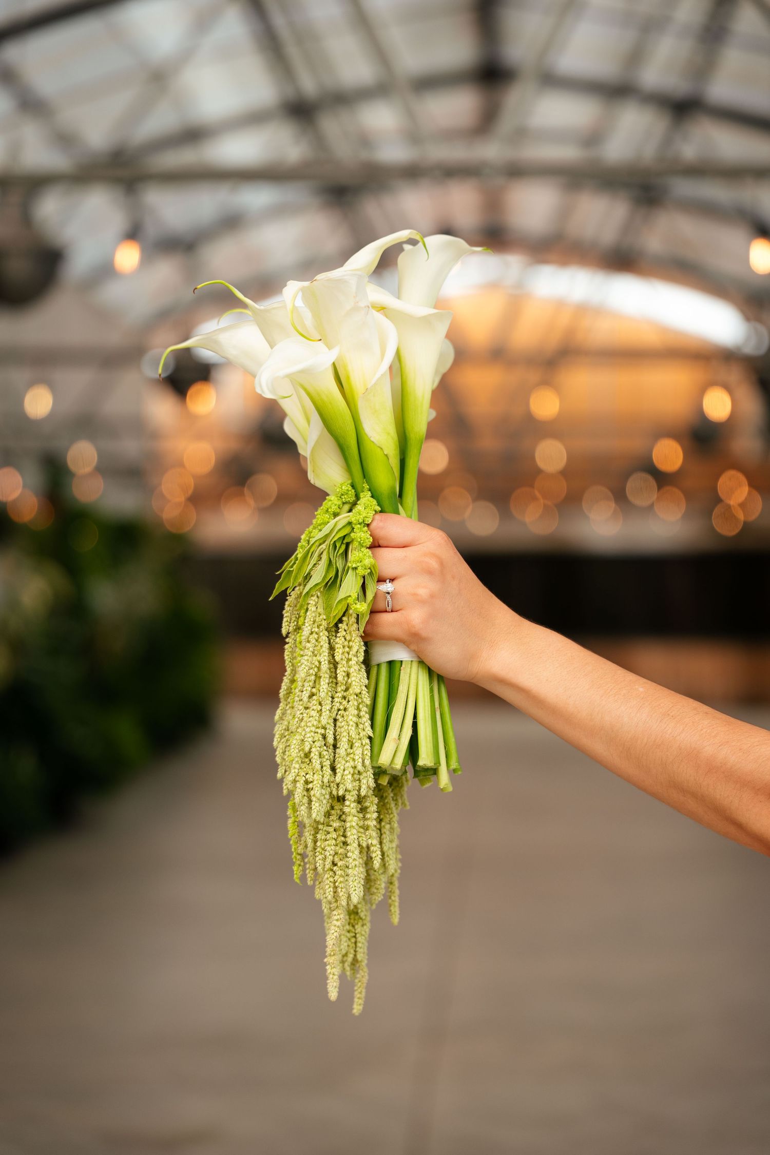 Hand holding elegant white calla lily bouquet with green amaranthus in a warmly lit greenhouse venue.