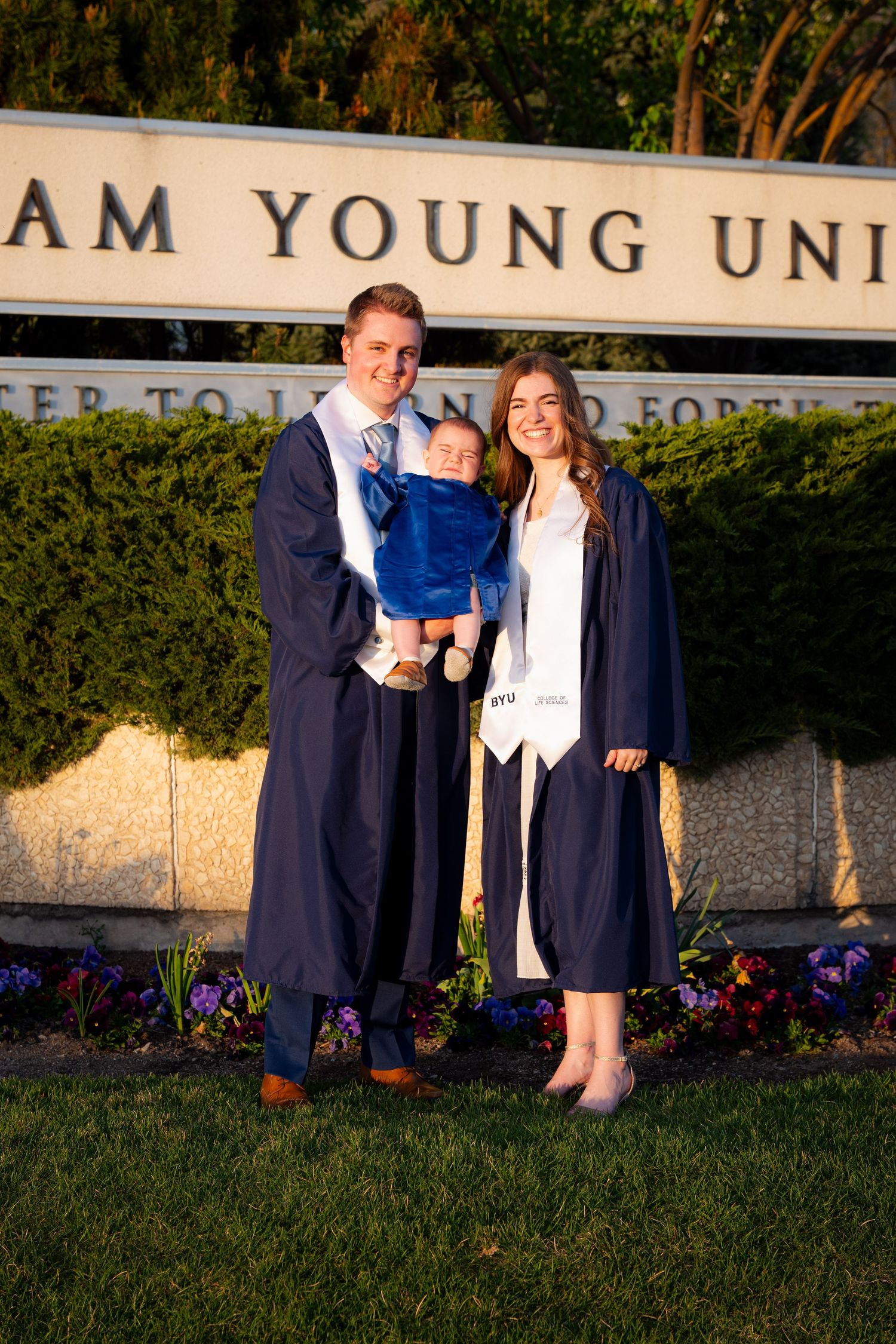 A family of three, including a young child, stands in front of a sign for Brigham Young University.
