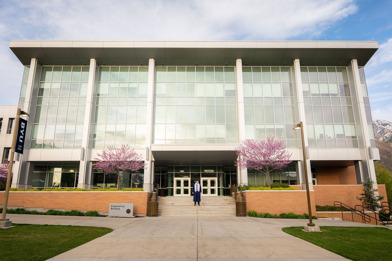 A man in a graduation gown is standing in front of a building with red flowers.