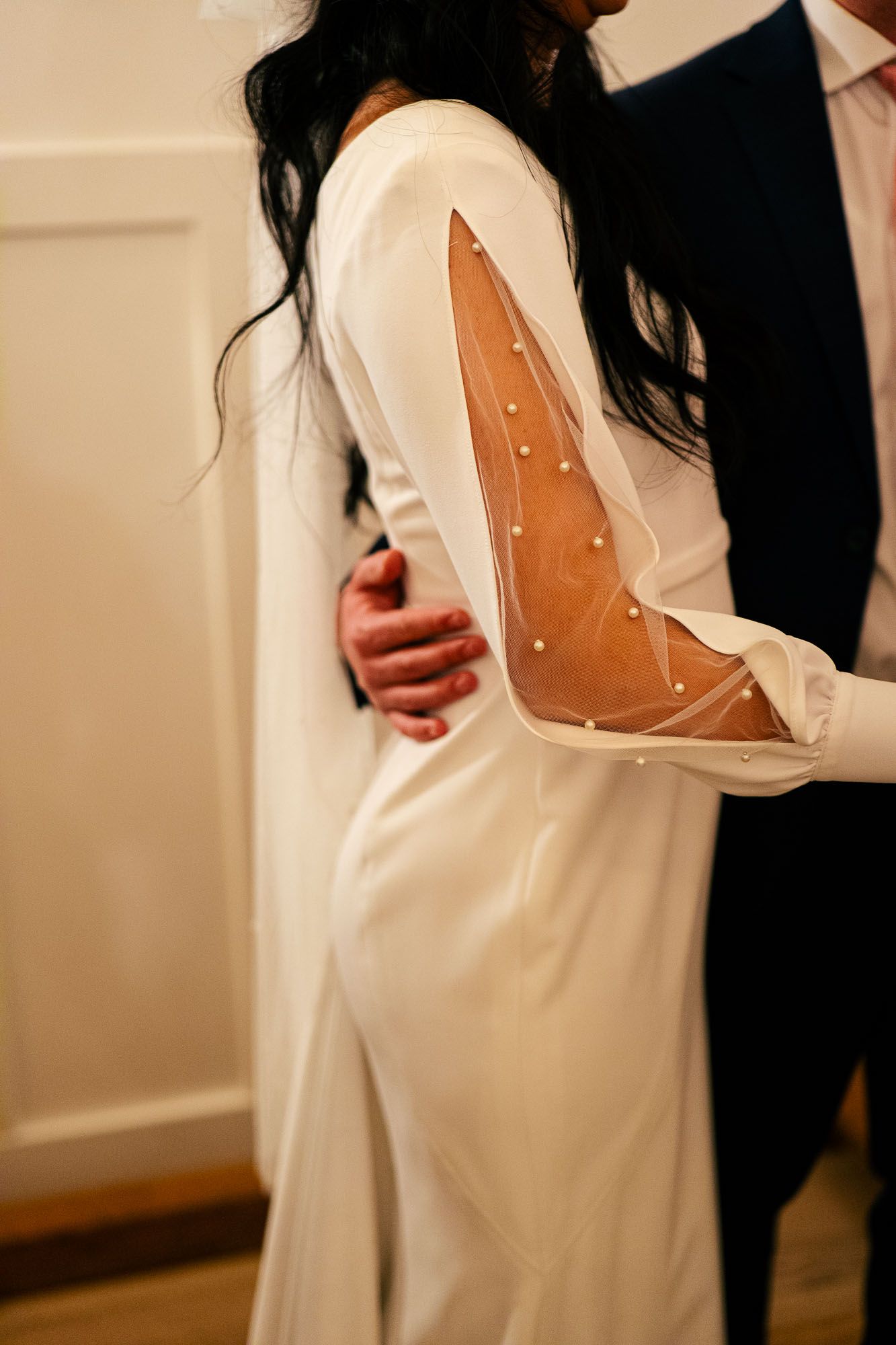 Bride in elegant white wedding gown with sheer pearl-embellished sleeves, embraced by groom in navy suit.