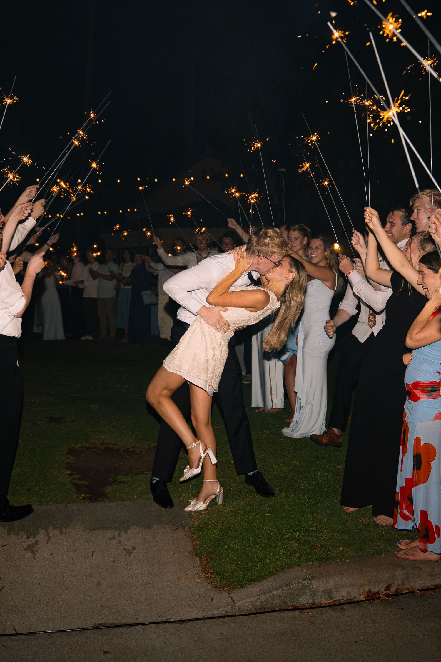Couple shares romantic dip kiss during sparkler send-off at outdoor wedding reception at night.