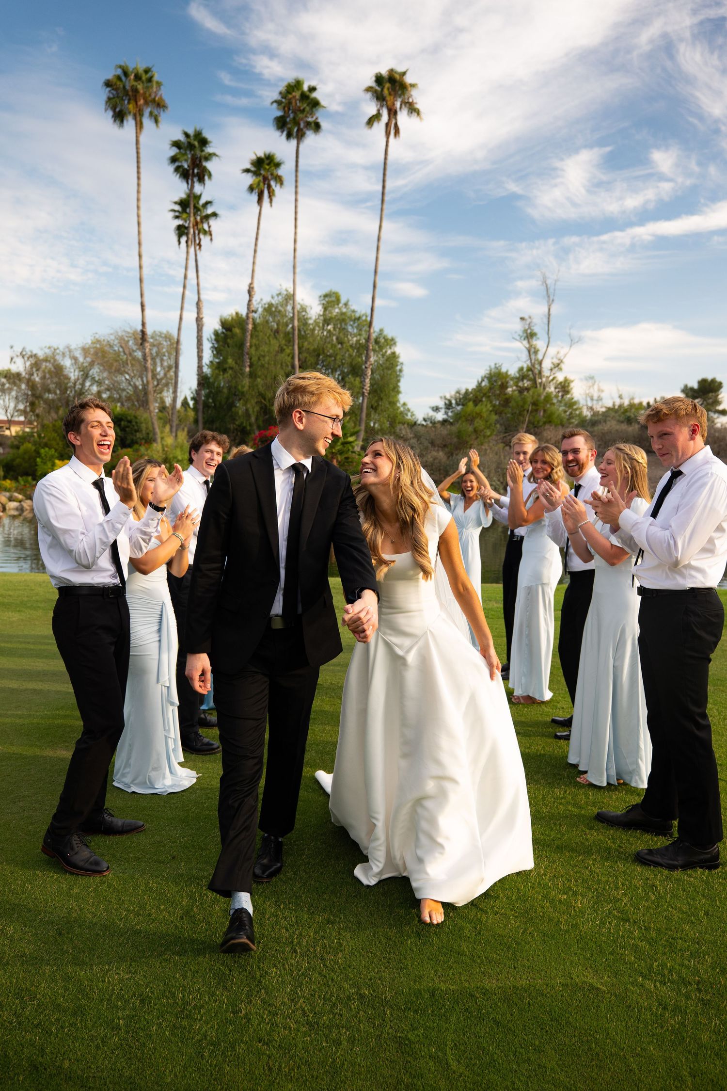 Bride and groom walk hand-in-hand through cheering wedding party on green lawn with palm trees backdrop.