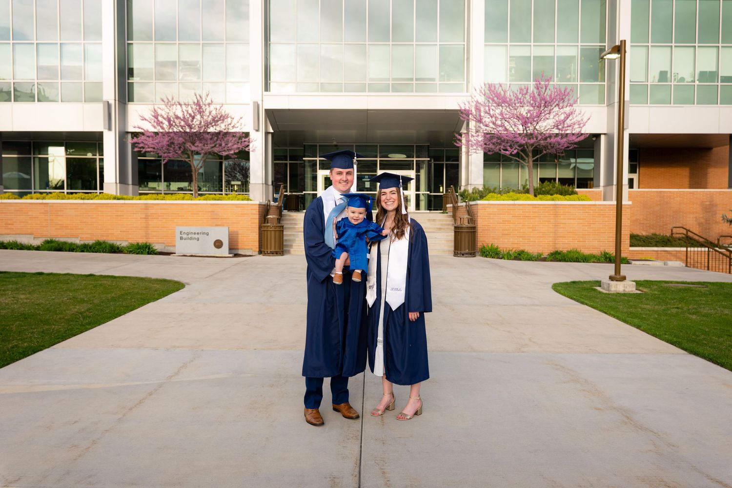 Two people wearing graduation gowns and caps stand together in front of a university building.