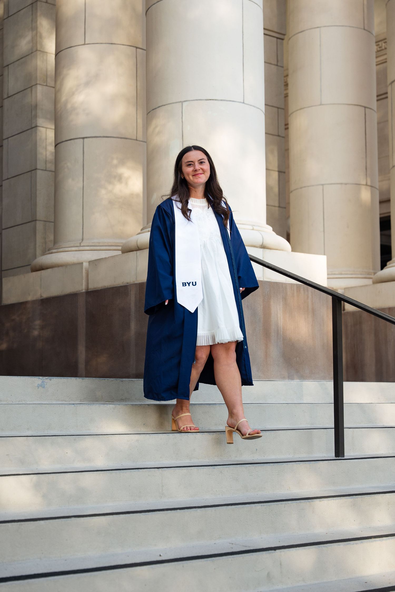 BYU female graduate standing on stone steps beside columns in navy commencement gown and stole.