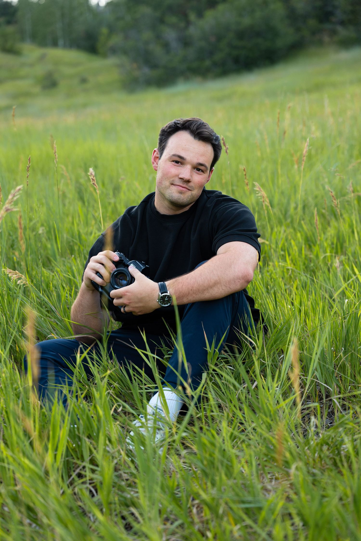 Man sitting in tall green grass holding a DSLR camera, wearing a black shirt and jeans, smiling outdoors.