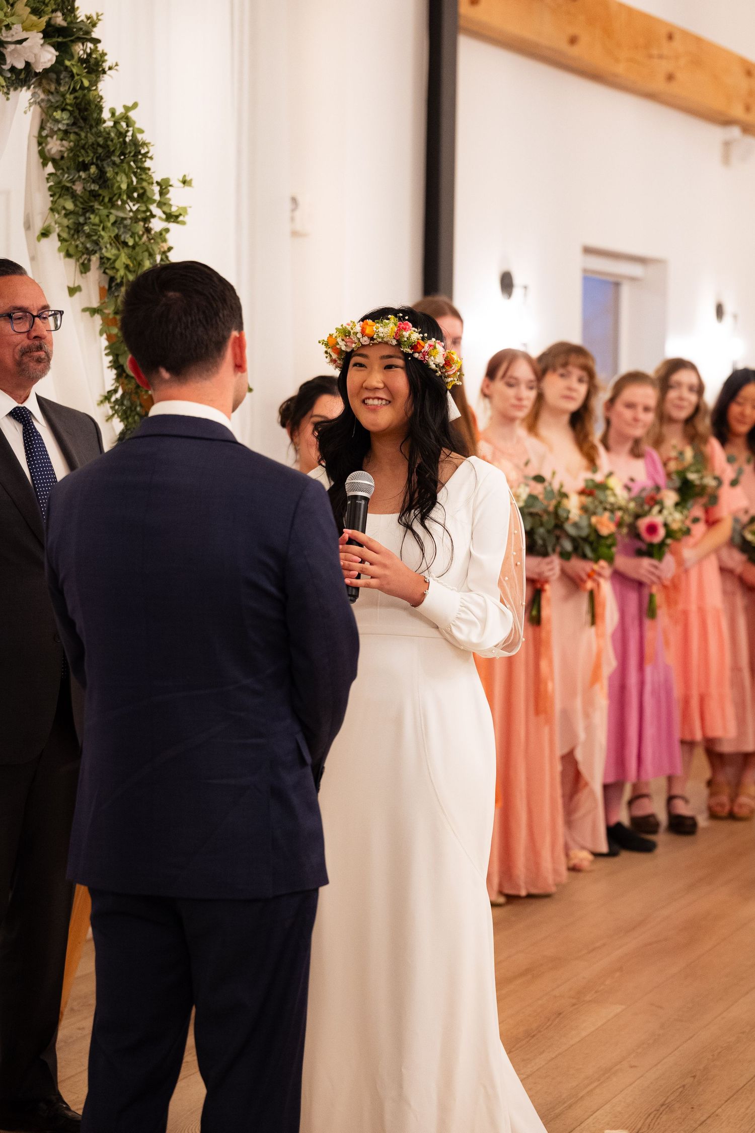 Smiling bride in white dress and colorful floral crown recites vows into microphone at elegant indoor wedding ceremony.