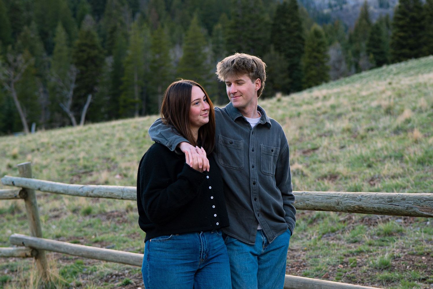 A young couple smiles together near a wooden fence with green forested hills in the background.