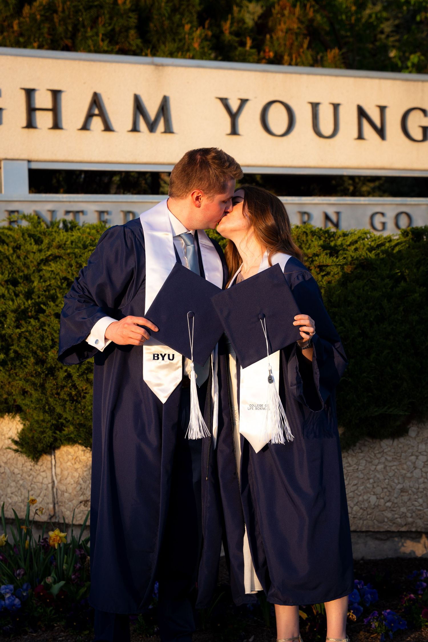 A woman in a graduation gown and cap stands in front of a university building.
