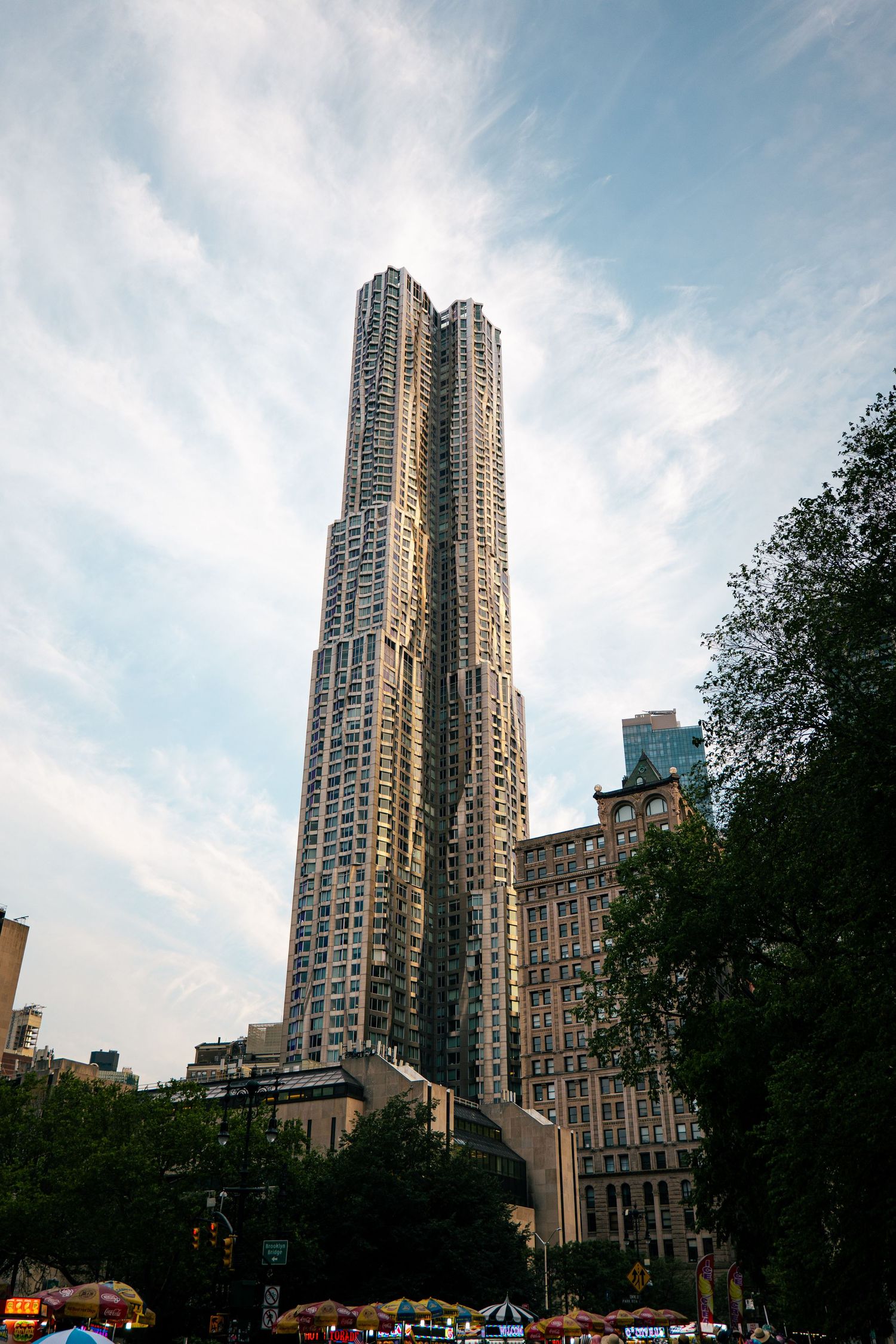 The image shows a tall, modern skyscraper with a distinctive architectural design, set against a blue sky with wispy clouds.
