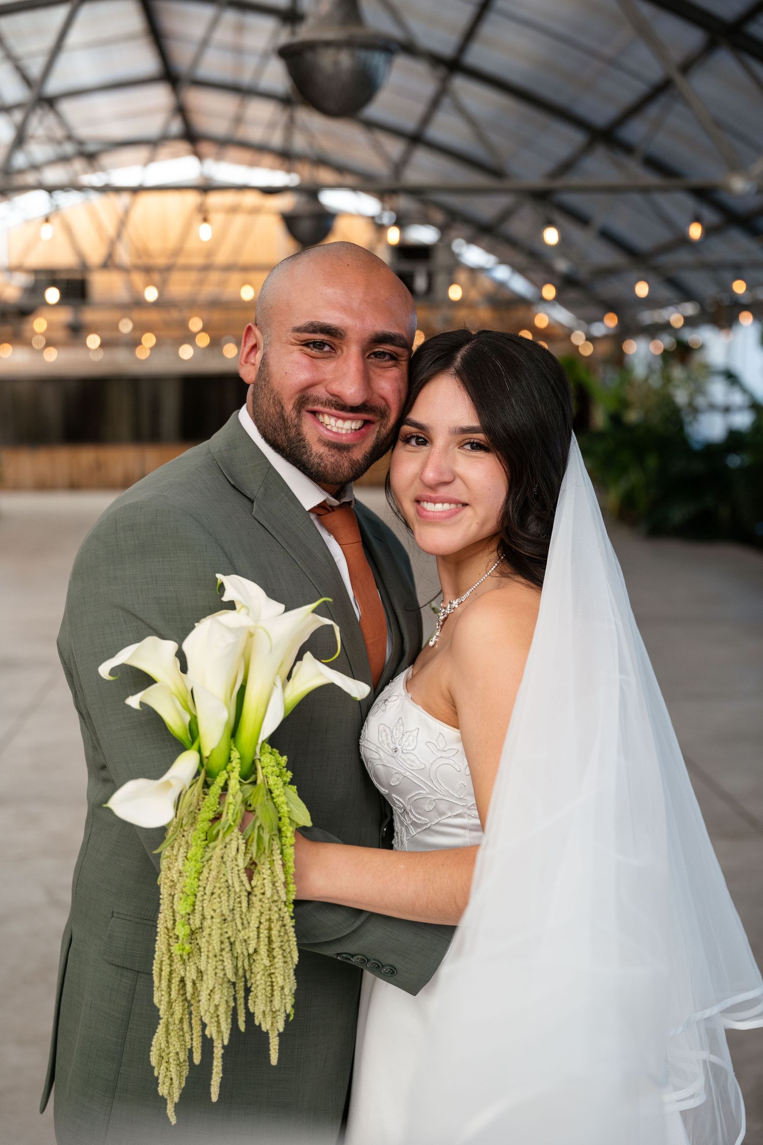Smiling bride and groom posing with white calla lily bouquet under string lights at elegant indoor wedding venue.