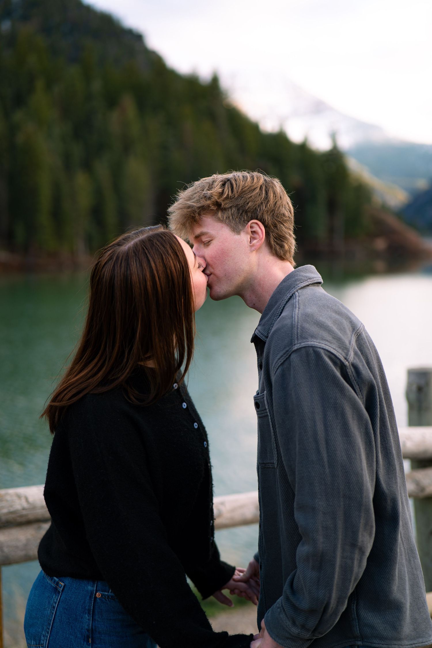 Couple sharing a romantic kiss on a wooden bridge with a scenic mountain lake and forest backdrop.
