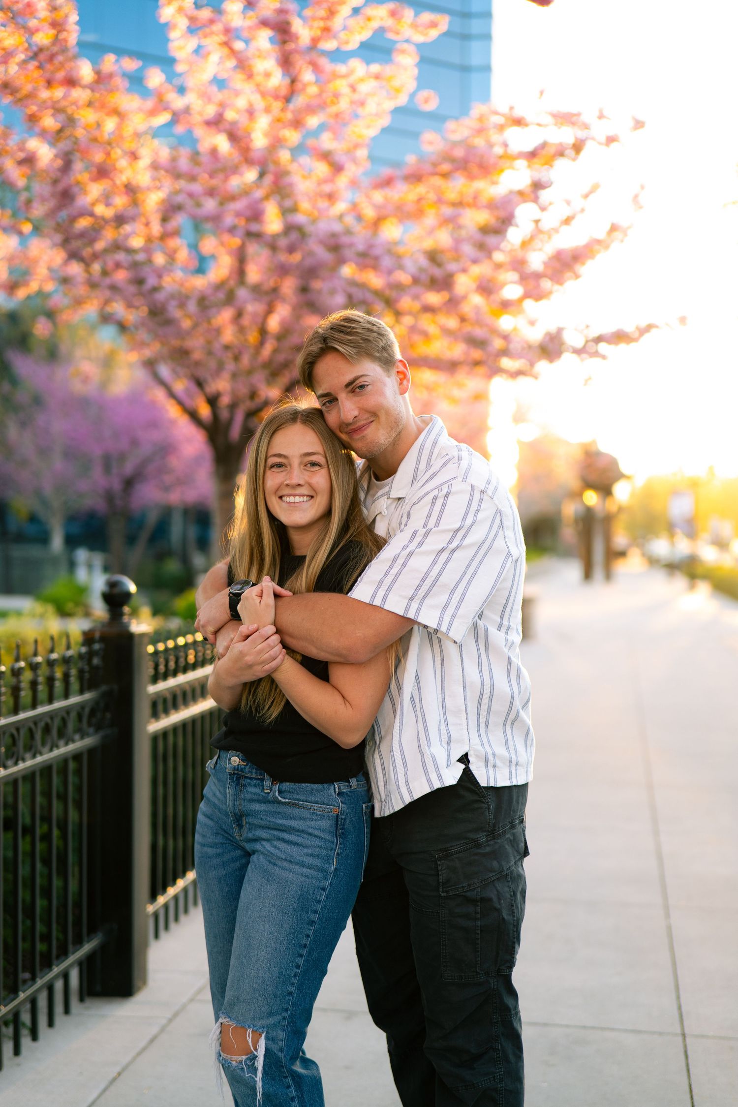 A young couple embraces and smiles outdoors near blooming cherry blossom trees at golden hour.