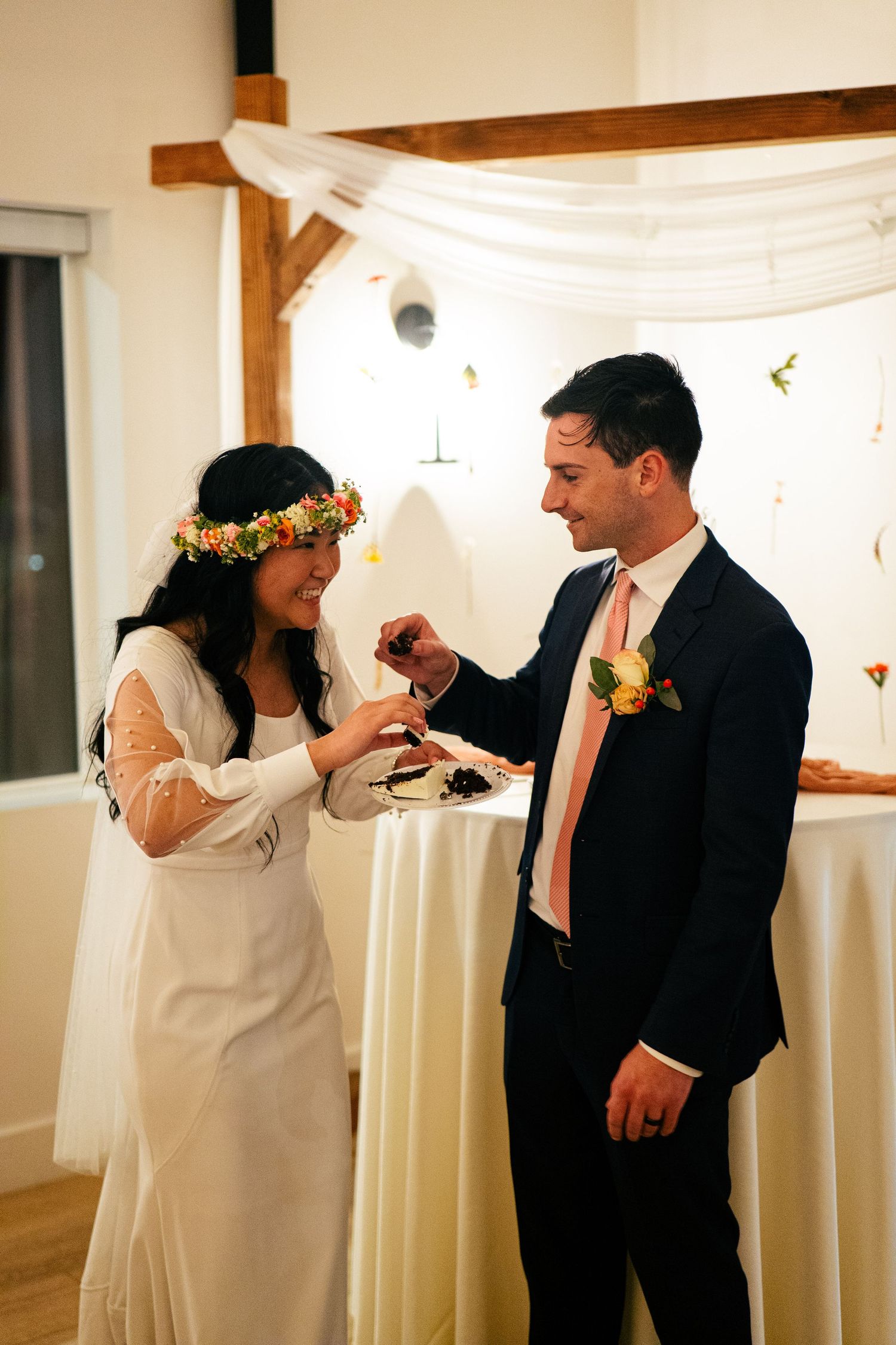 Bride in floral crown and groom in navy suit sharing wedding cake during reception celebration.