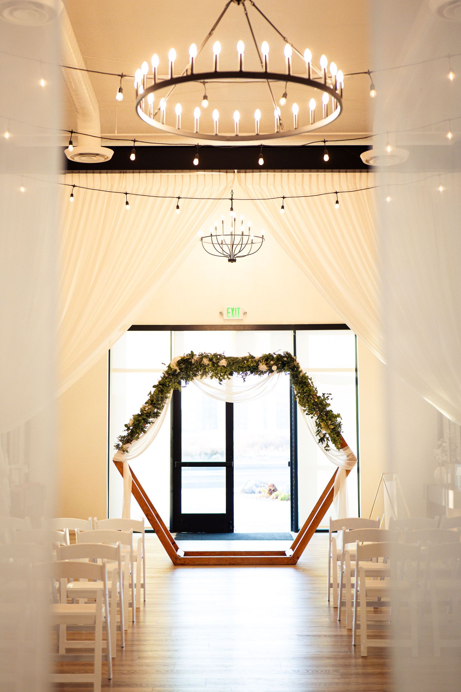Elegant wedding ceremony venue with hexagonal floral arch, white chairs, draped fabric, and chandelier lighting.