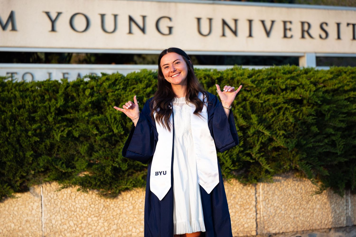 Graduate in cap and gown poses proudly in front of BYU sign, flashing school spirit hand gestures.