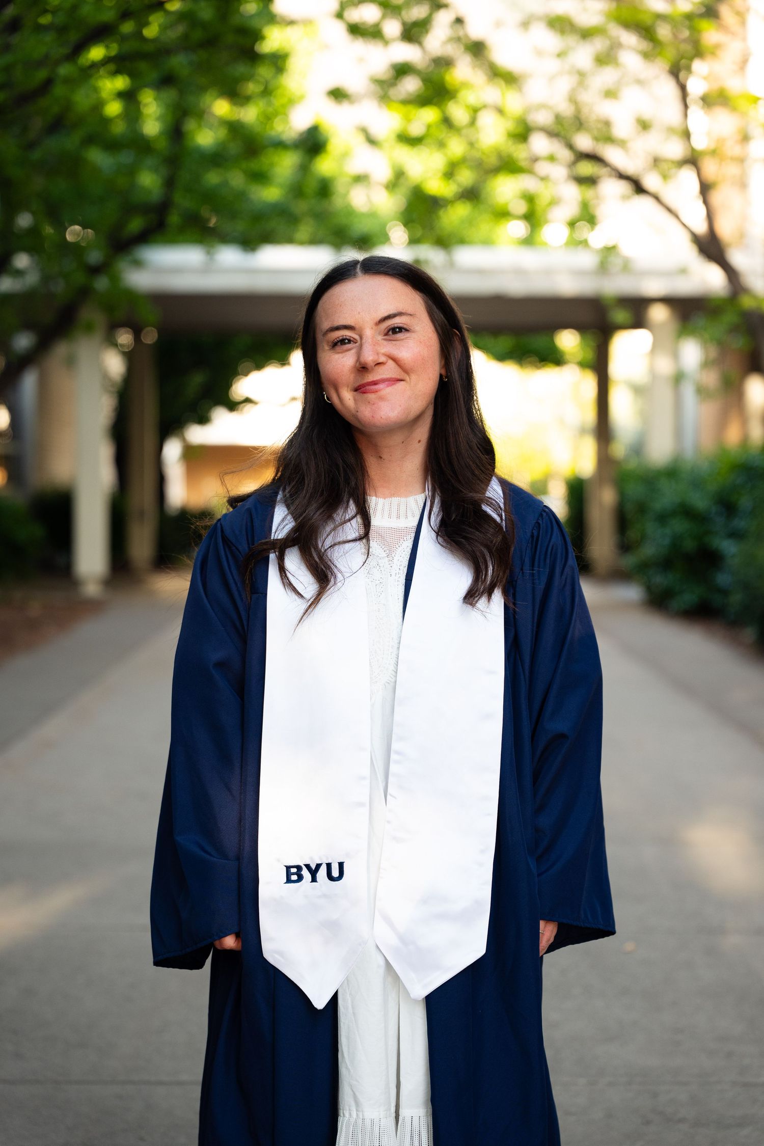 BYU graduate smiling in navy cap and gown with white BYU stole on a tree-lined campus walkway.