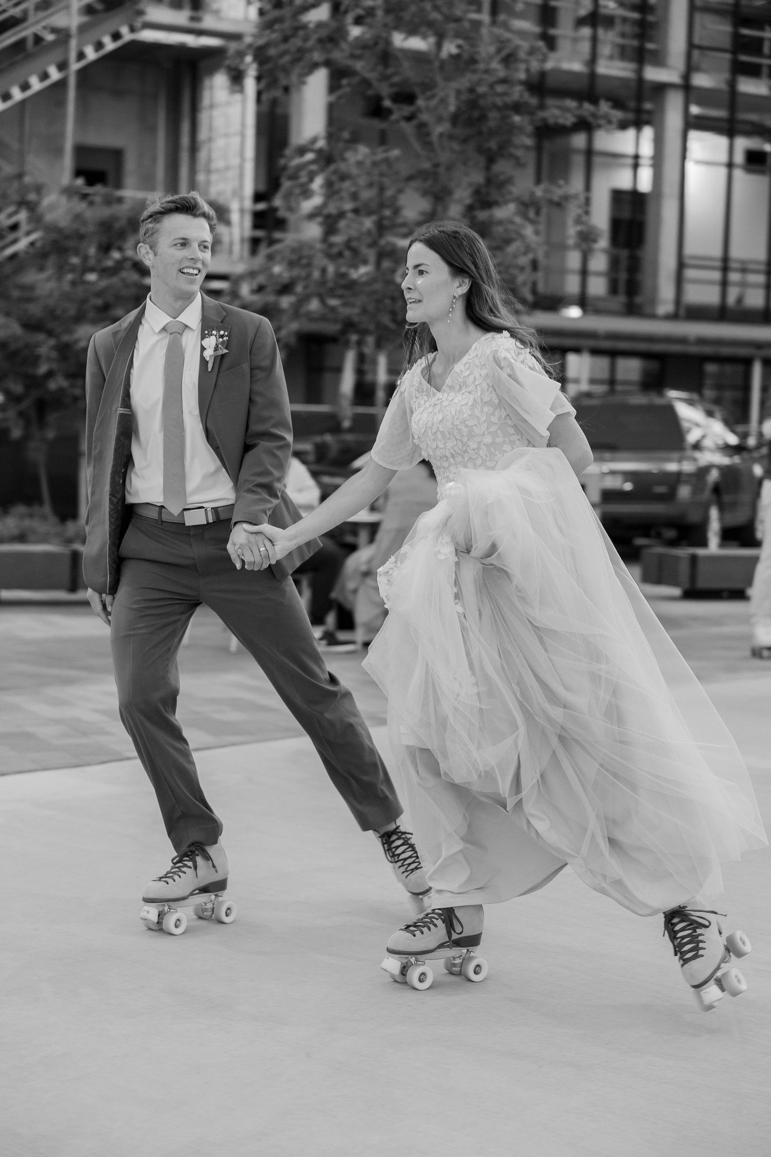 Black and white photo of a bride and groom holding hands while roller skating outdoors in wedding attire.
