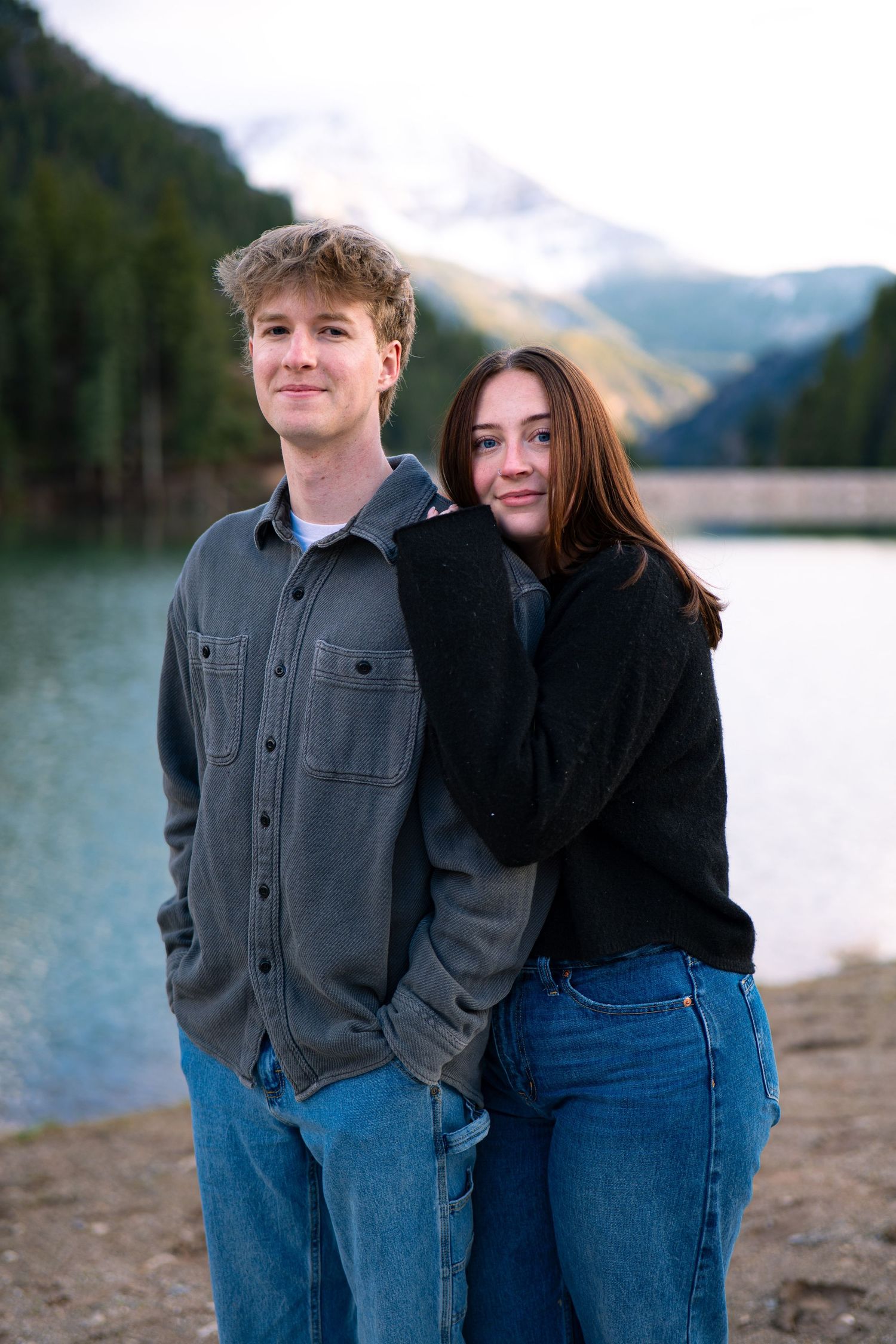 A young couple poses by a mountain lake with snow-capped peaks, wearing casual denim and sweaters.