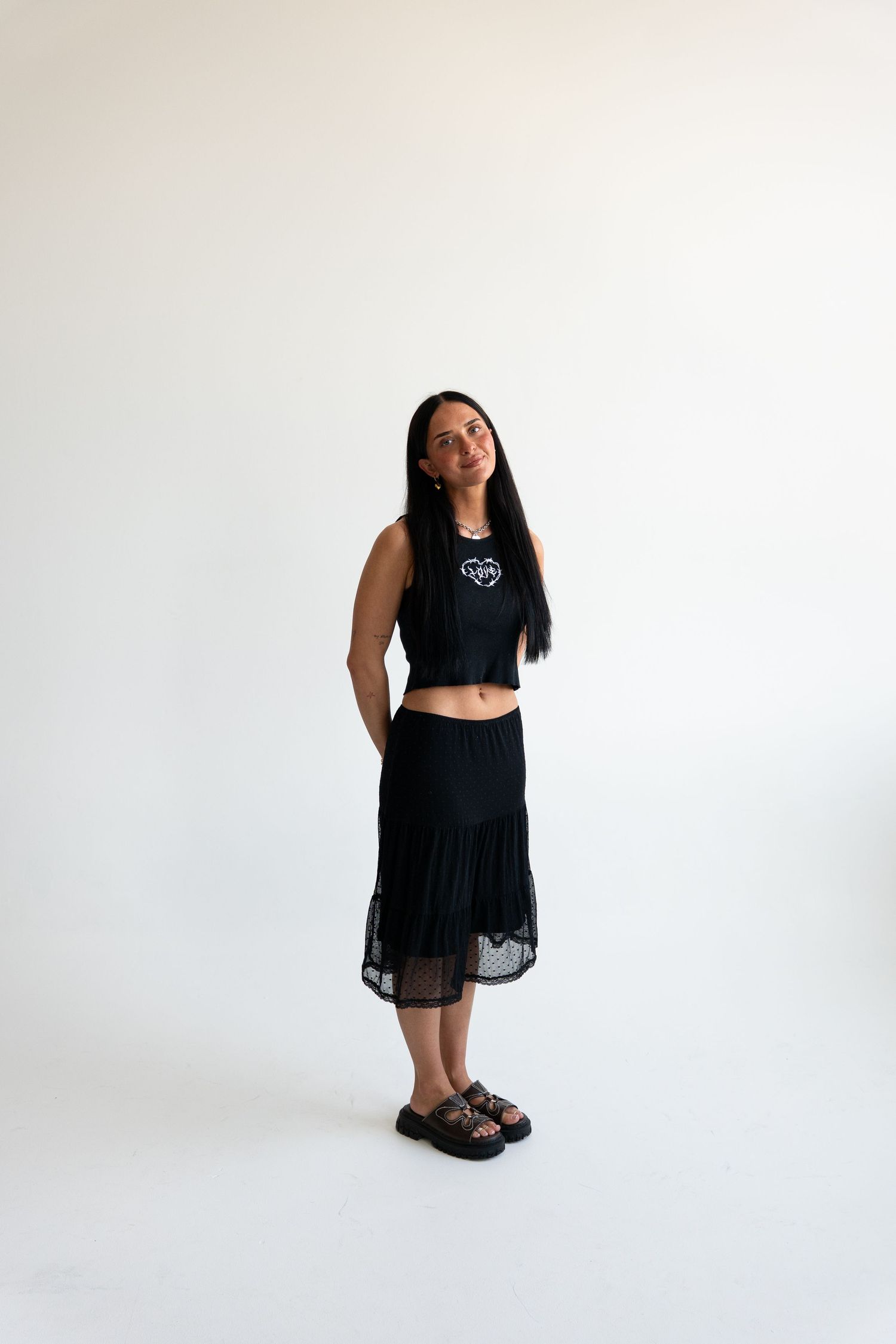 Woman in black crop top and midi lace skirt with leopard sandals posing against white background.