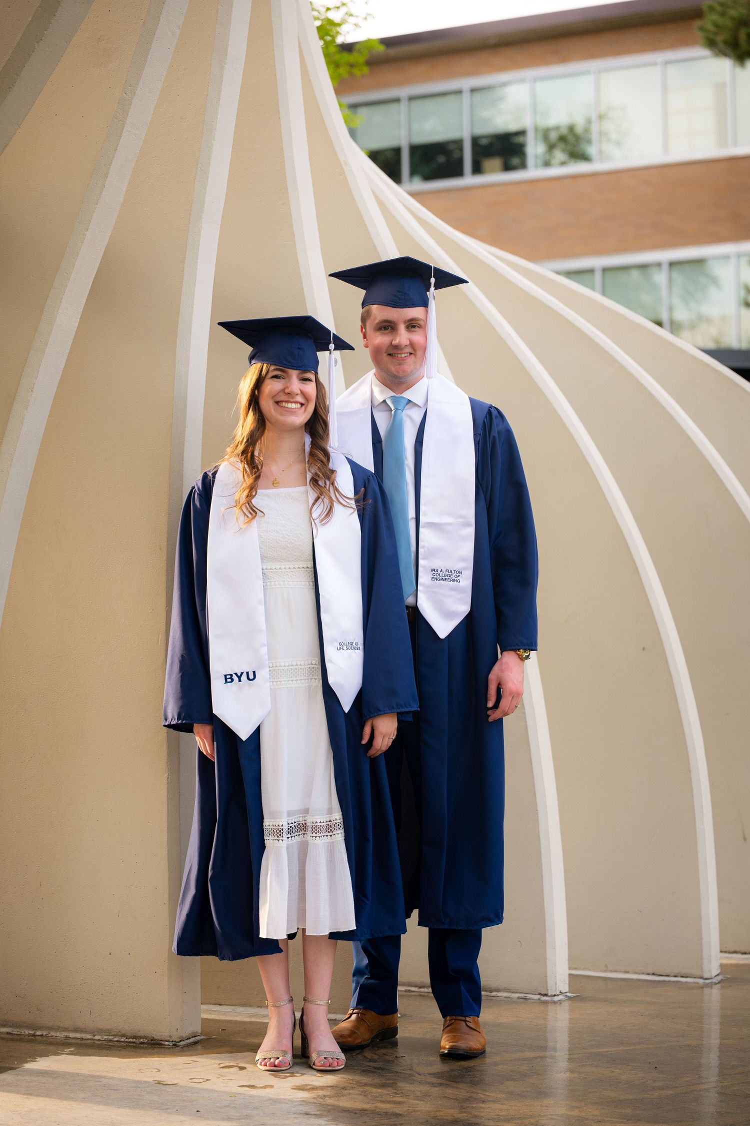 Two graduates in academic regalia stand together in front of a building, celebrating their achievement.