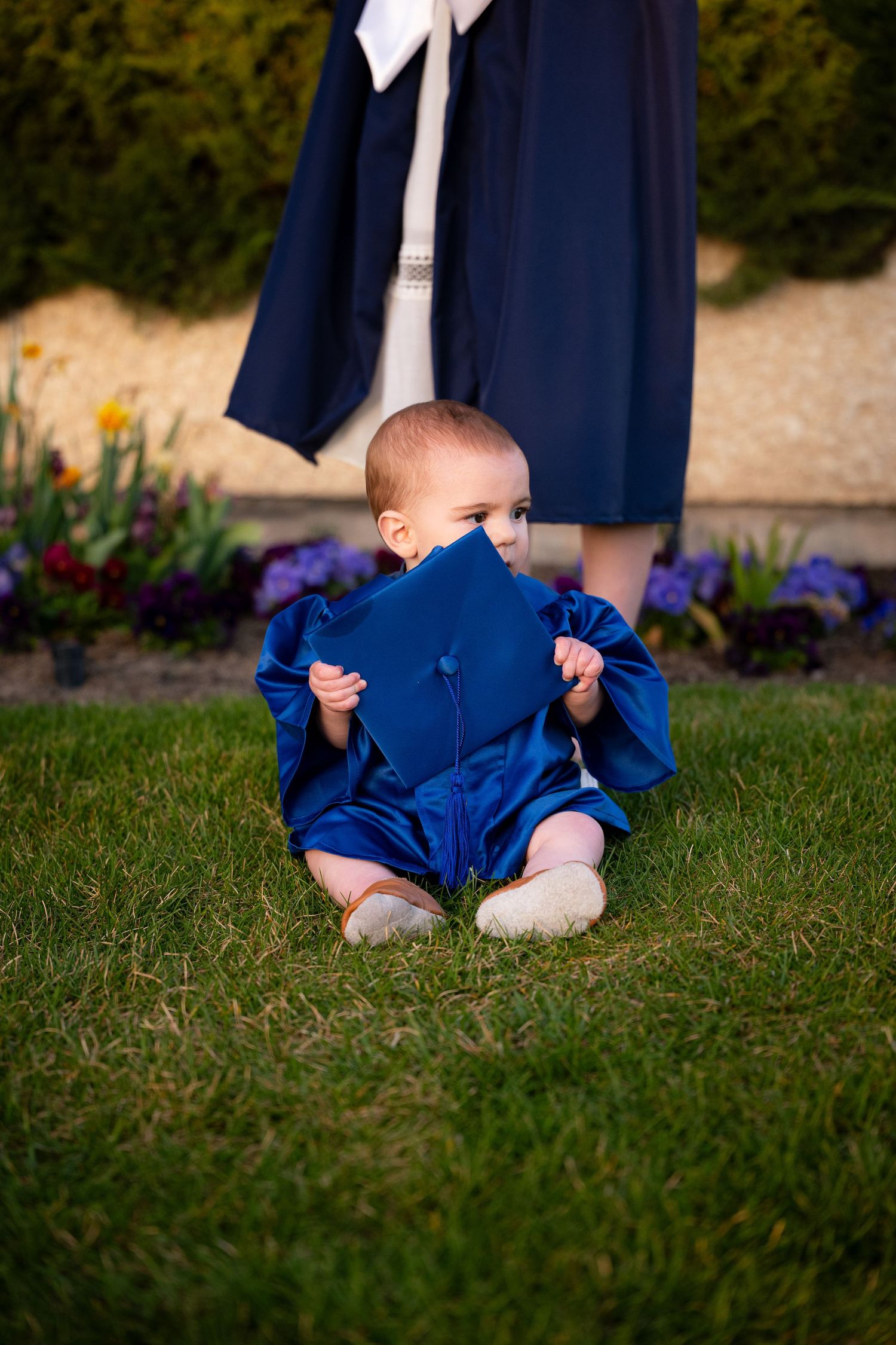A baby is sitting on the grass in a blue outfit, with a woman in a navy blue dress standing behind them.