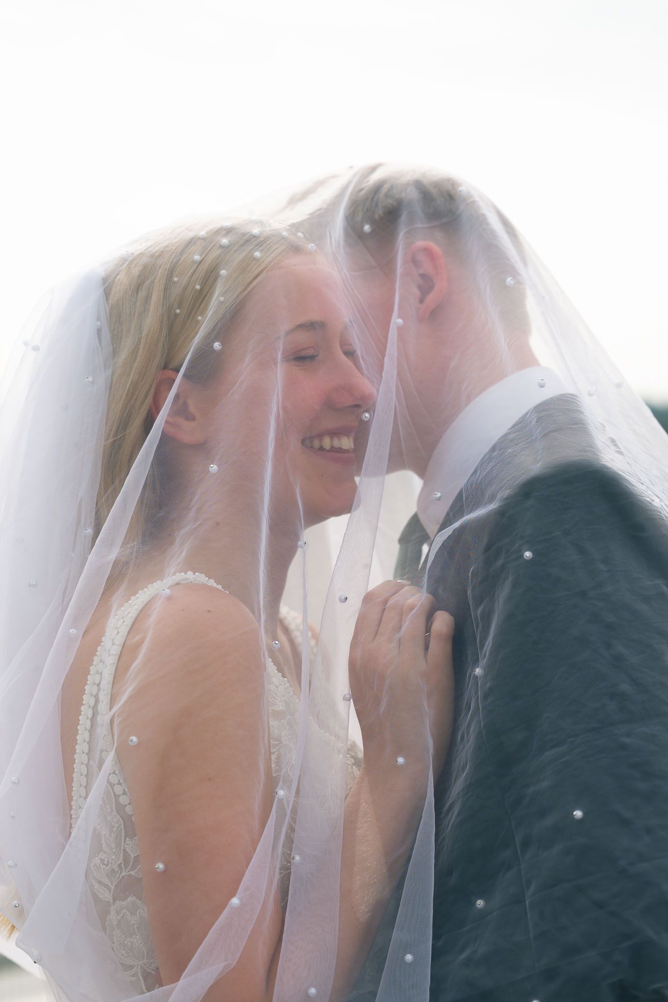 Bride and groom share a romantic moment under a pearl-adorned wedding veil, smiling together on their wedding day.