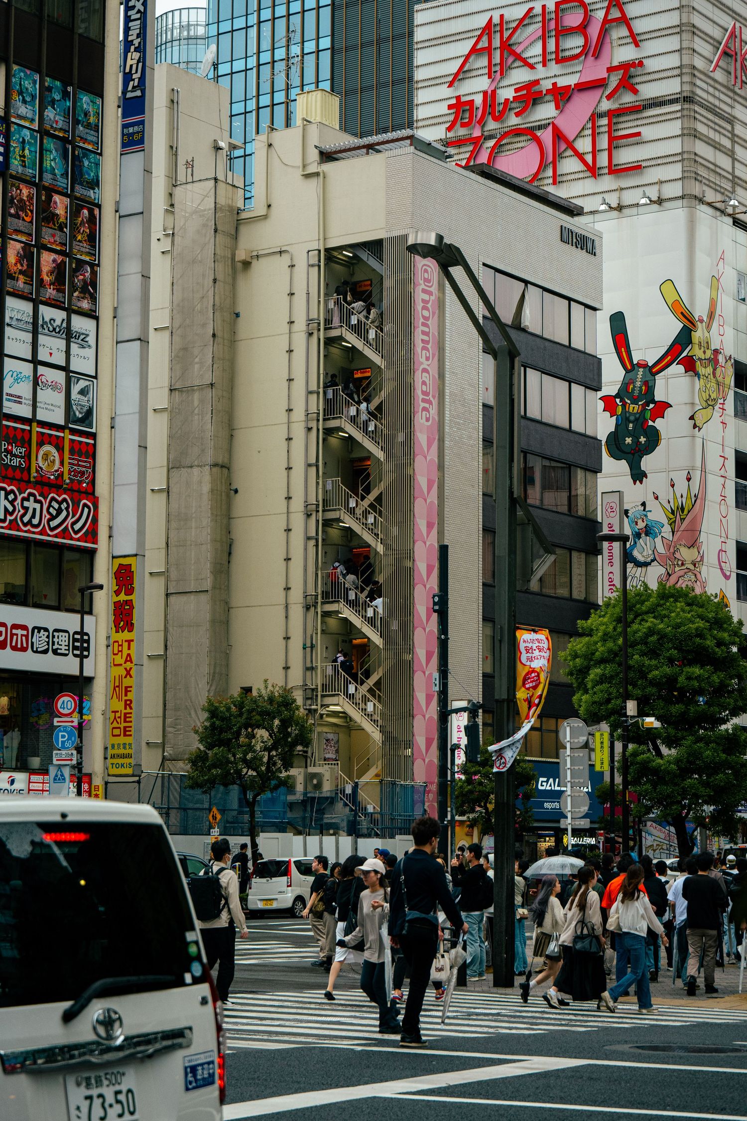 A crowded city street in Japan, with a large Akihabara sign and various shops and businesses visible.