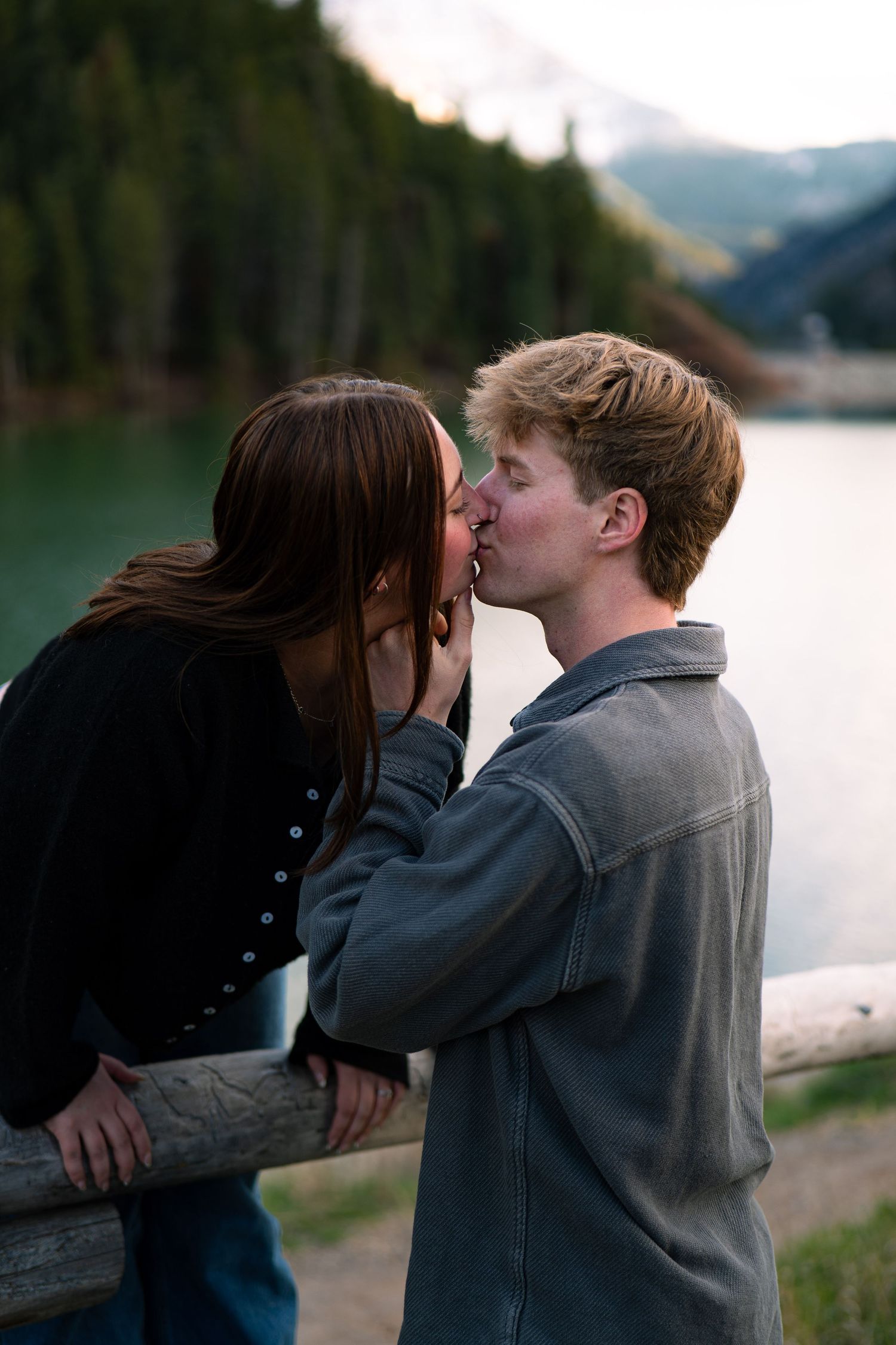 A young couple shares a romantic kiss by a serene mountain lake surrounded by evergreen trees at dusk.