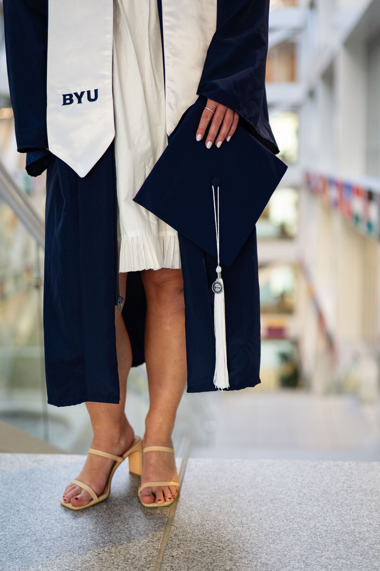 BYU graduate in navy cap and gown holding mortarboard, wearing gold heels in university hallway.