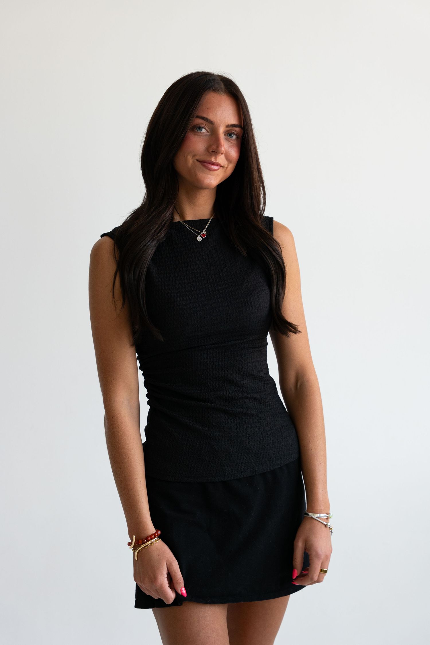 Tan brunette woman in black sleeveless ribbed top and mini skirt smiling in a white studio setting.