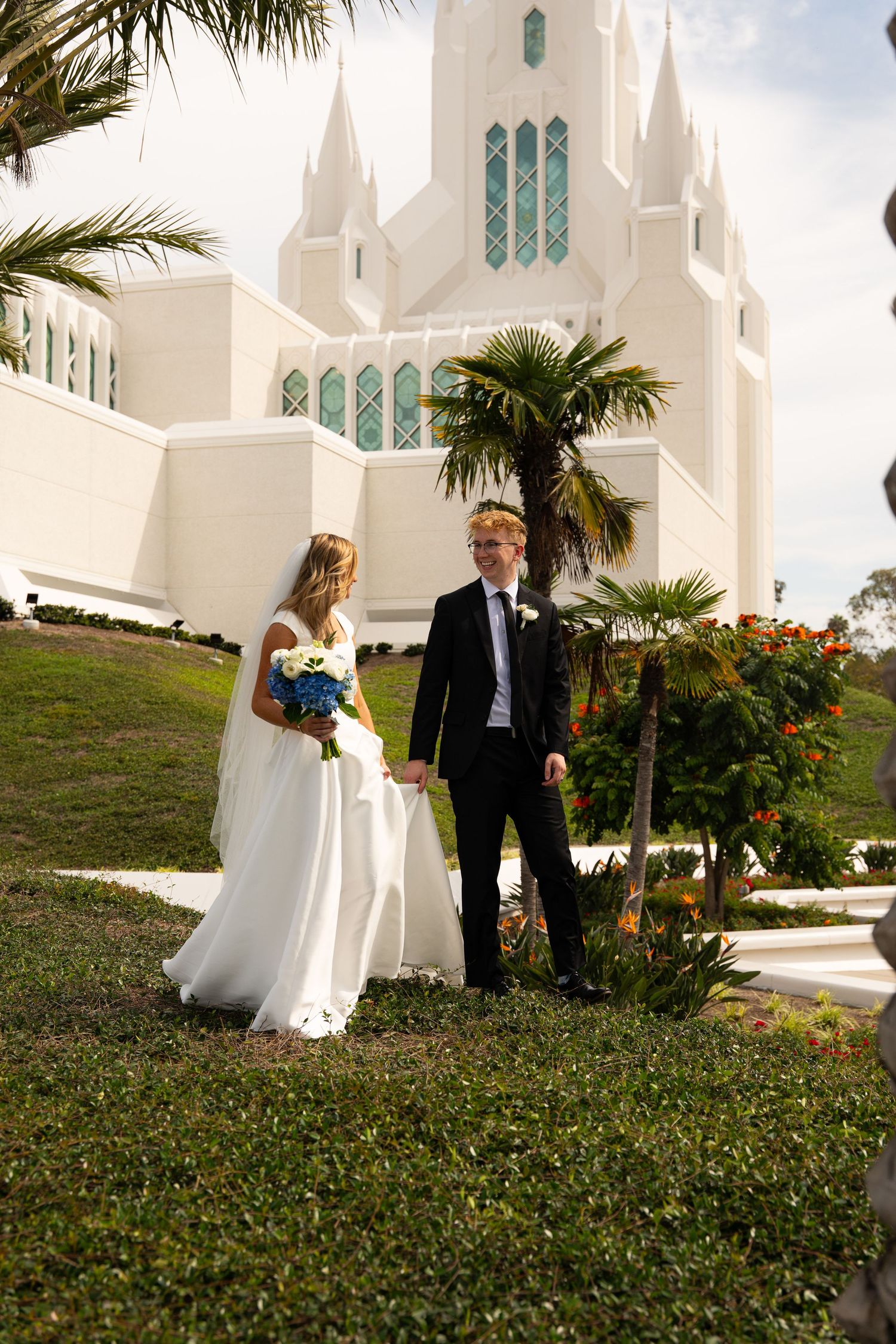 Bride and groom holding hands outside white San Diego LDS Temple surrounded by tropical palm trees and gardens.