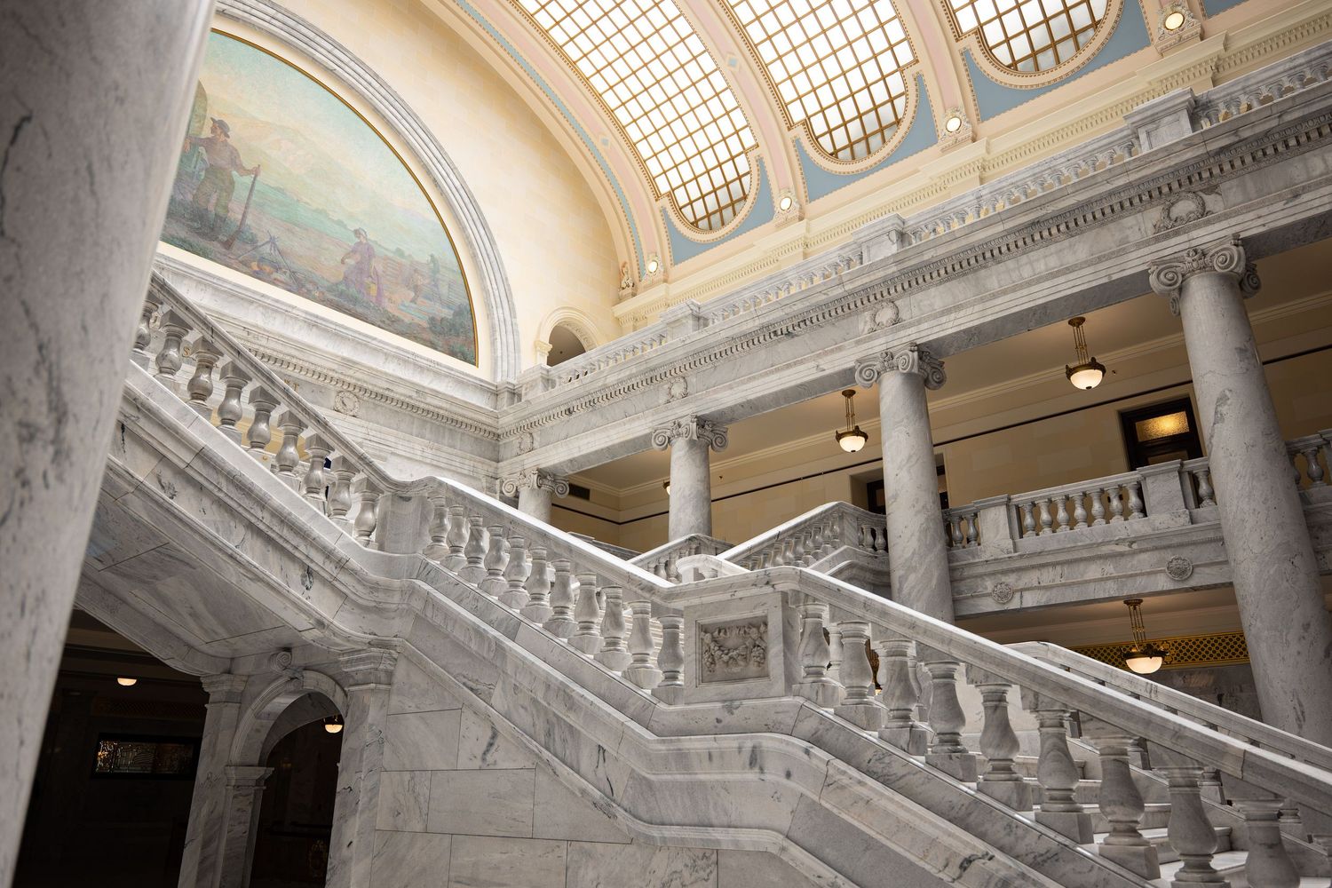 Grand marble staircase inside a government capitol building with ornate columns and a decorative painted mural.