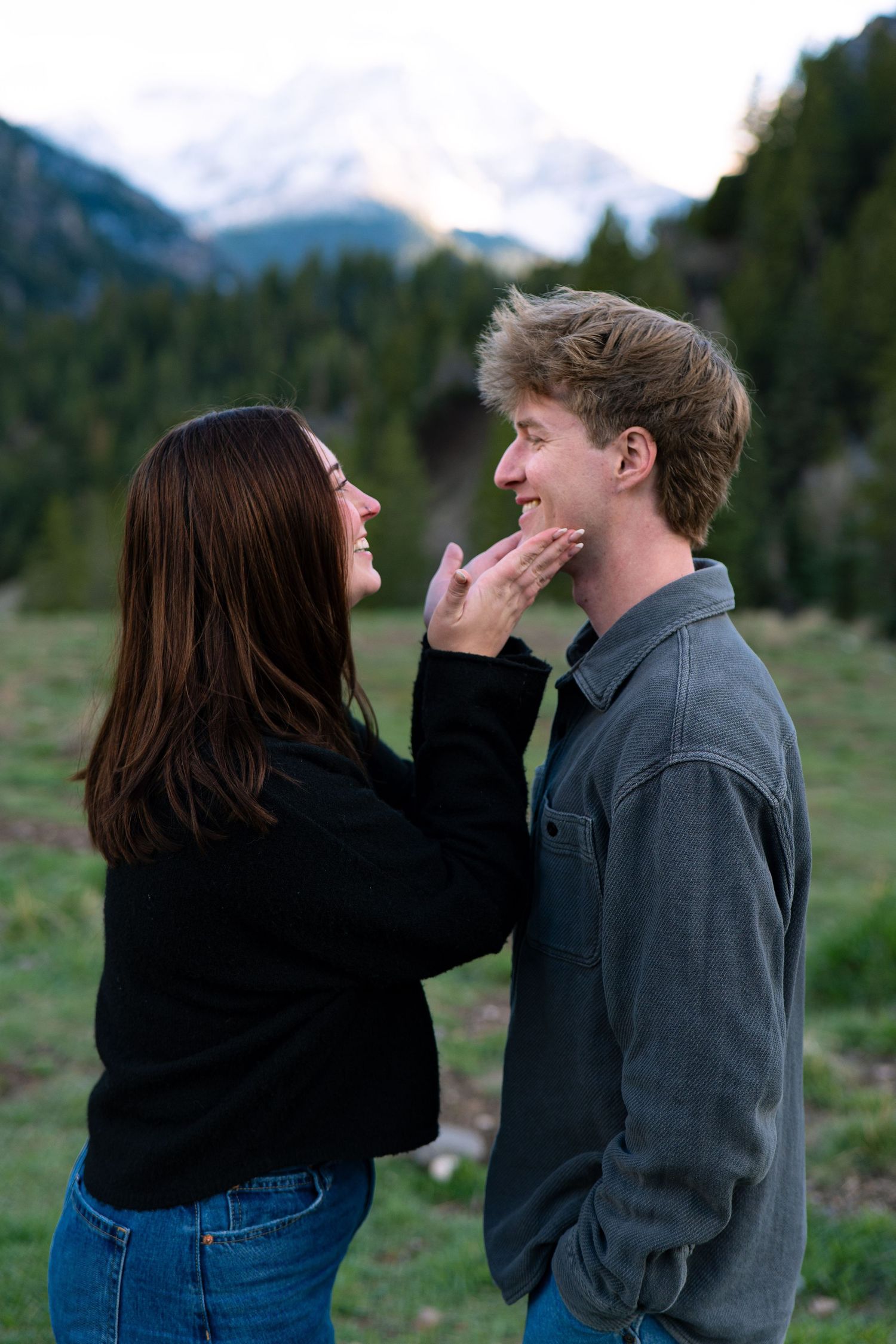 Couple sharing a playful moment in a mountain meadow, woman touching man's face while both smile.
