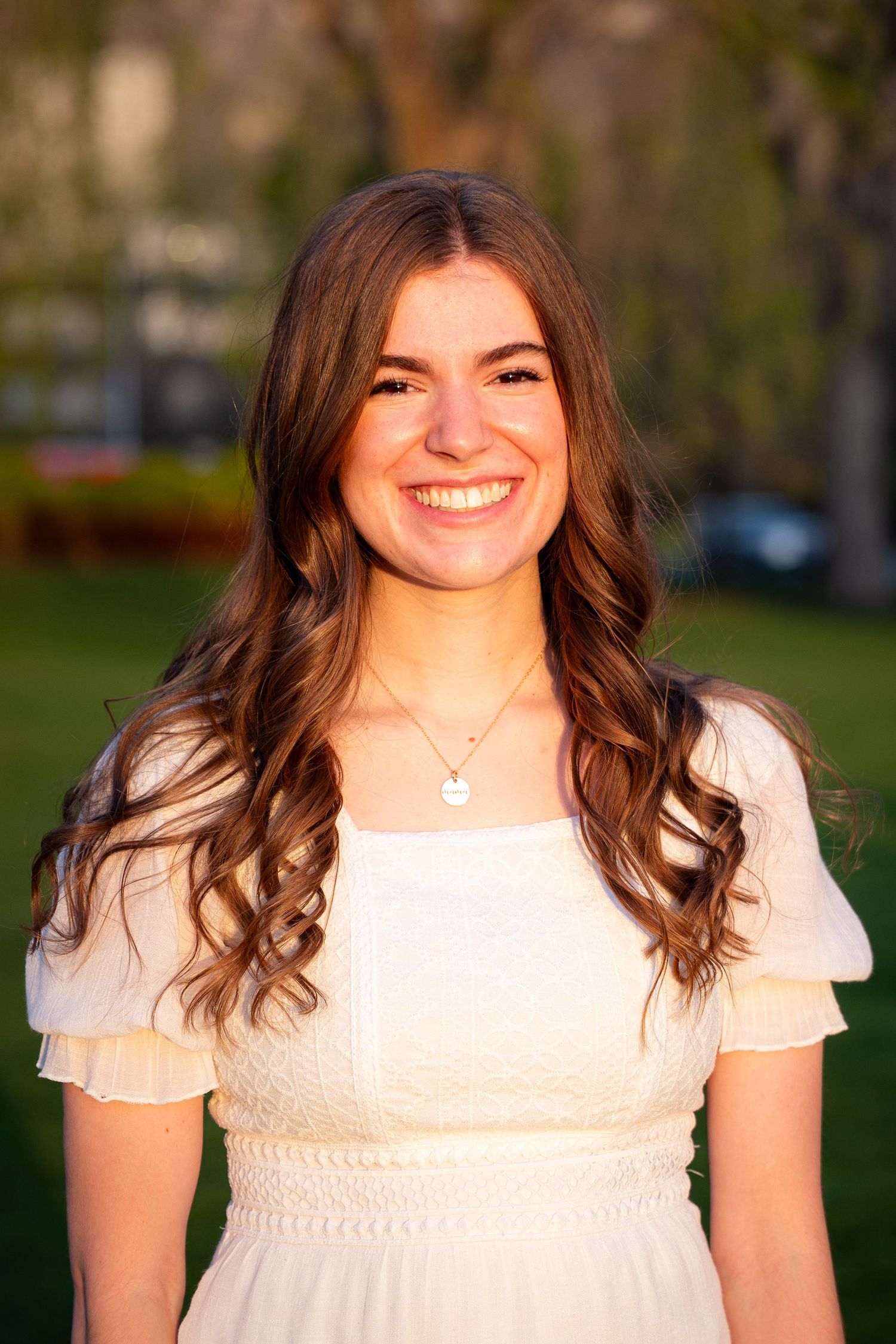 A young woman with long curly brown hair smiles while wearing a white dress in a grassy outdoor setting.