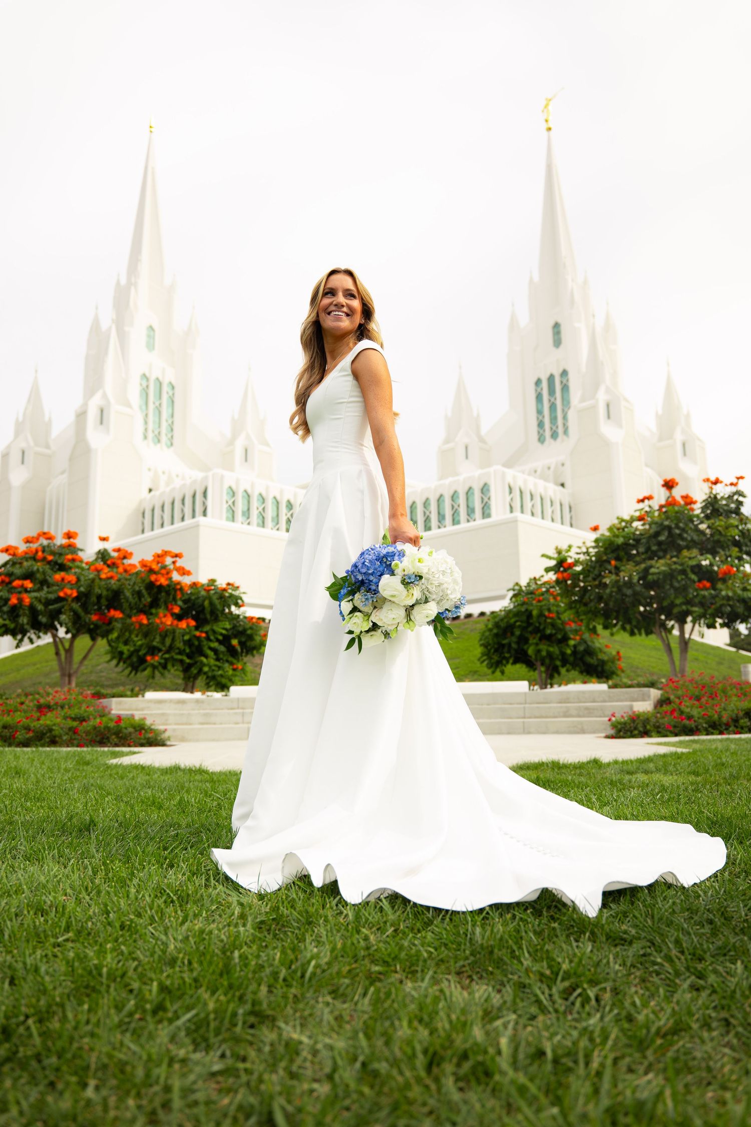 Bride in white gown holding blue and white bouquet stands before a white LDS temple with orange flowers.