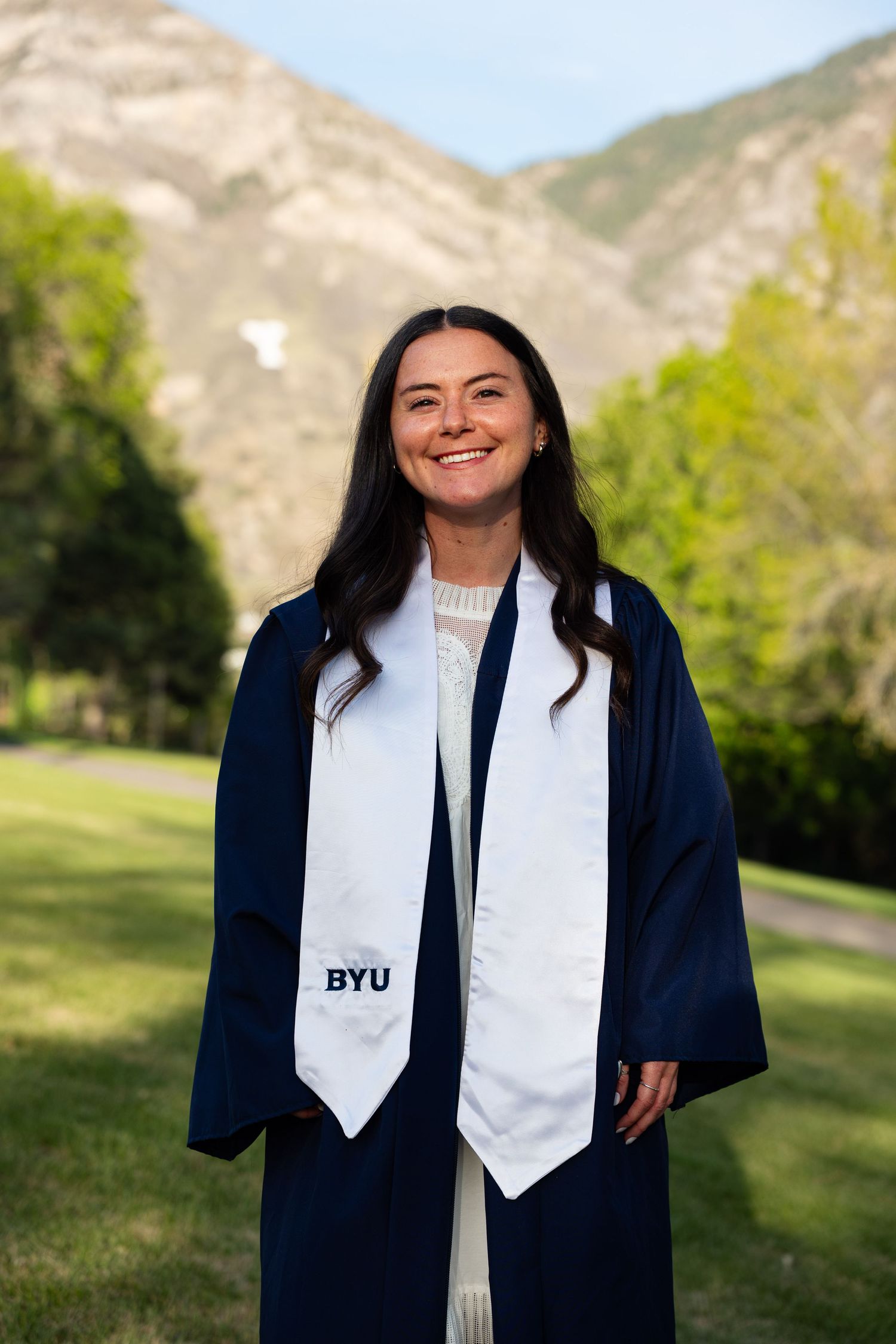 Smiling BYU graduate in navy cap and gown with white stole, posing outdoors with mountain backdrop.