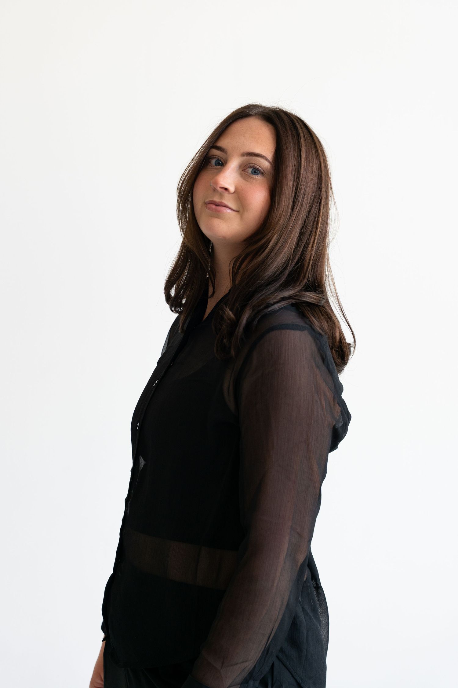 Brunette woman in a black sheer blouse posing sideways in a minimalist white studio setting.