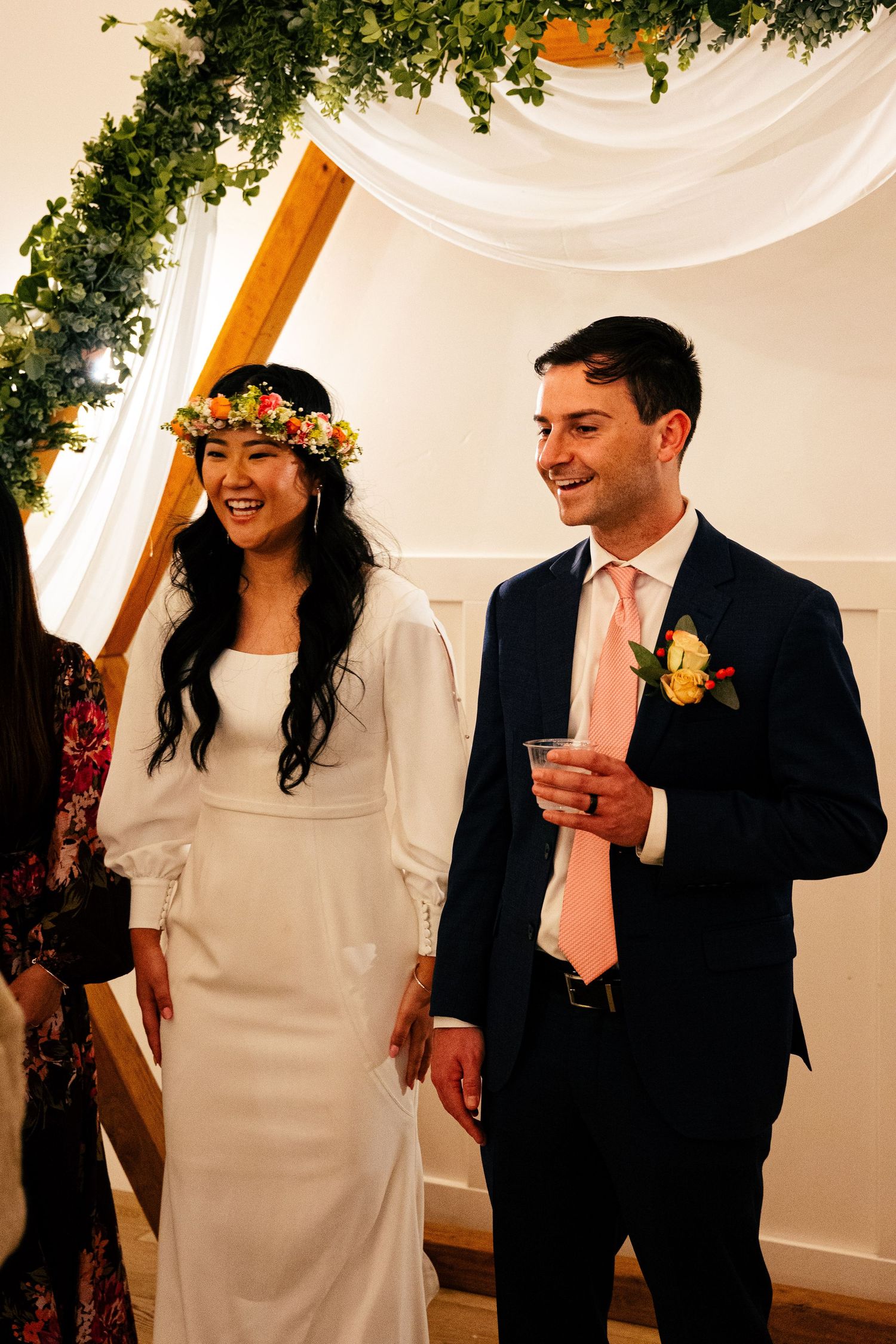 Smiling bride in white dress with floral crown and groom in navy suit with pink tie at wedding ceremony.