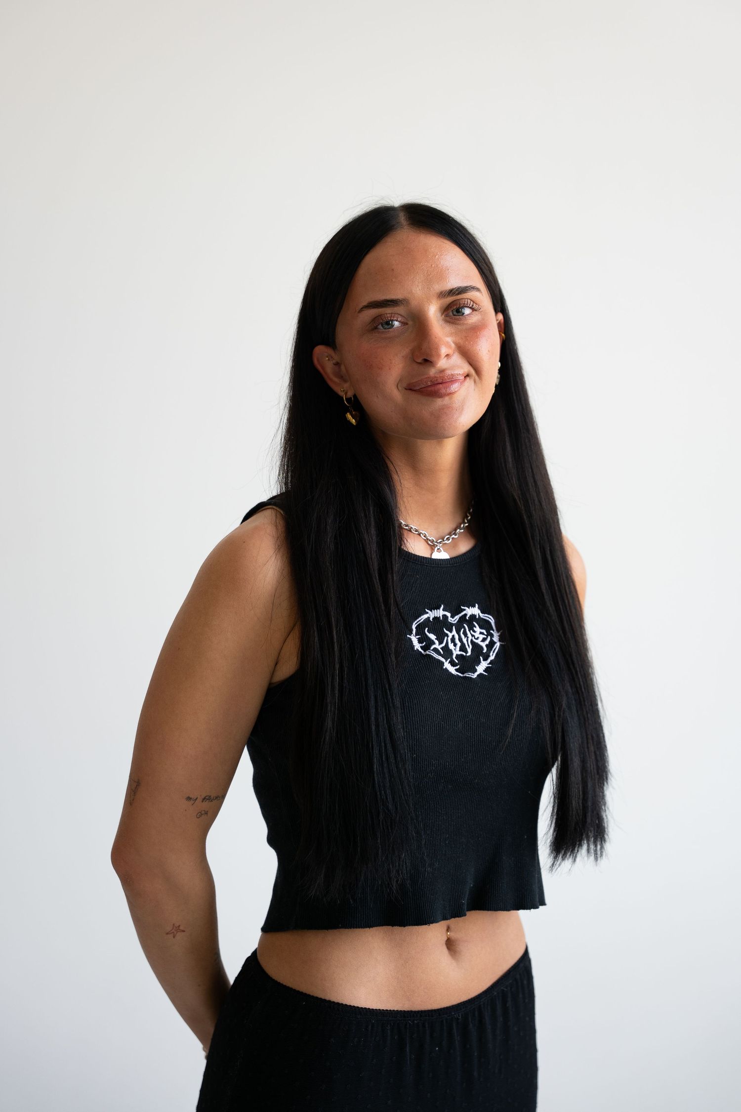 Woman with long black hair wearing a black crop tank top with heart graphic and black pants, smiling against white background.