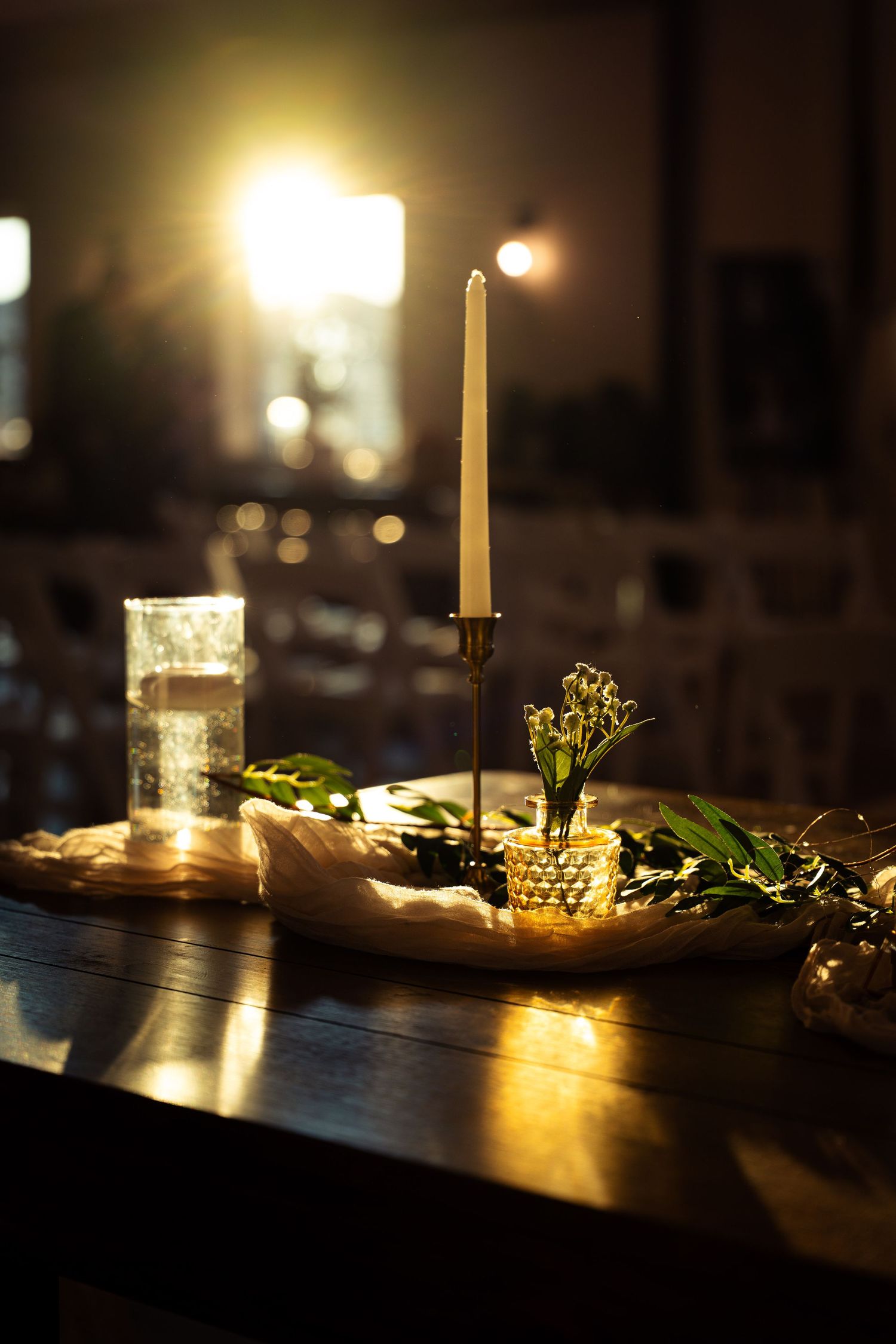 Elegant candlelit wedding table centerpiece with white taper candle, greenery, and gold accents on dark wood.