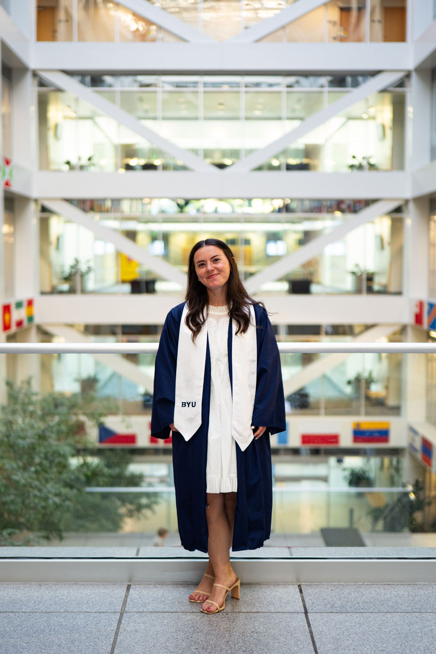 BYU graduate in navy cap and gown smiles in modern university atrium with white stole and heels.