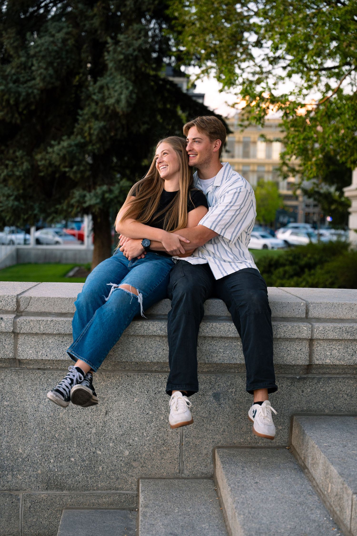 Young couple sitting on stone steps in a park, smiling and embracing on a warm sunny day.